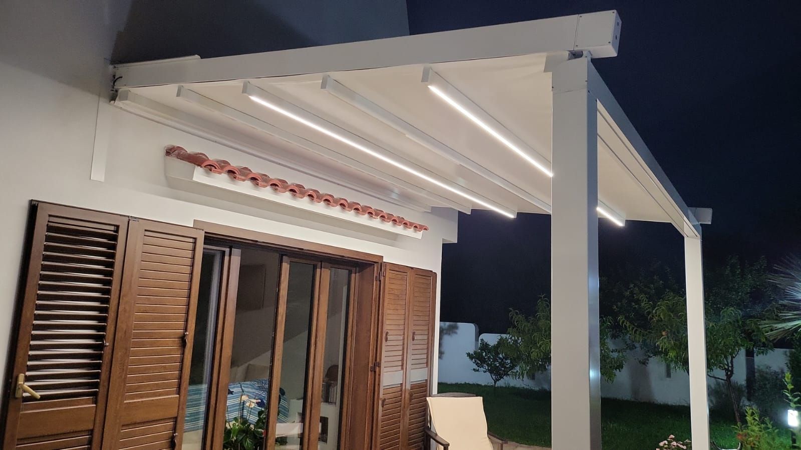 Pergola bianca con luci a LED sopra una porta a vetri e persiane in legno di notte.
