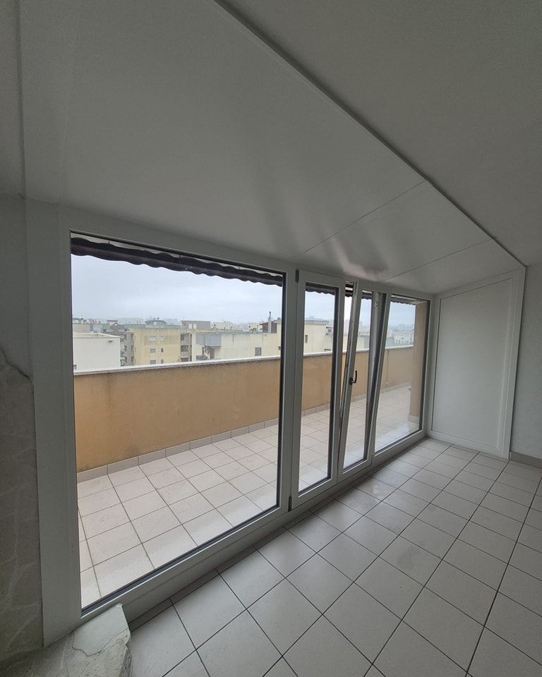 Vista interna della stanza con ampie finestre che si affacciano su un balcone e sul paesaggio urbano. Pavimento piastrellato, pareti bianche.