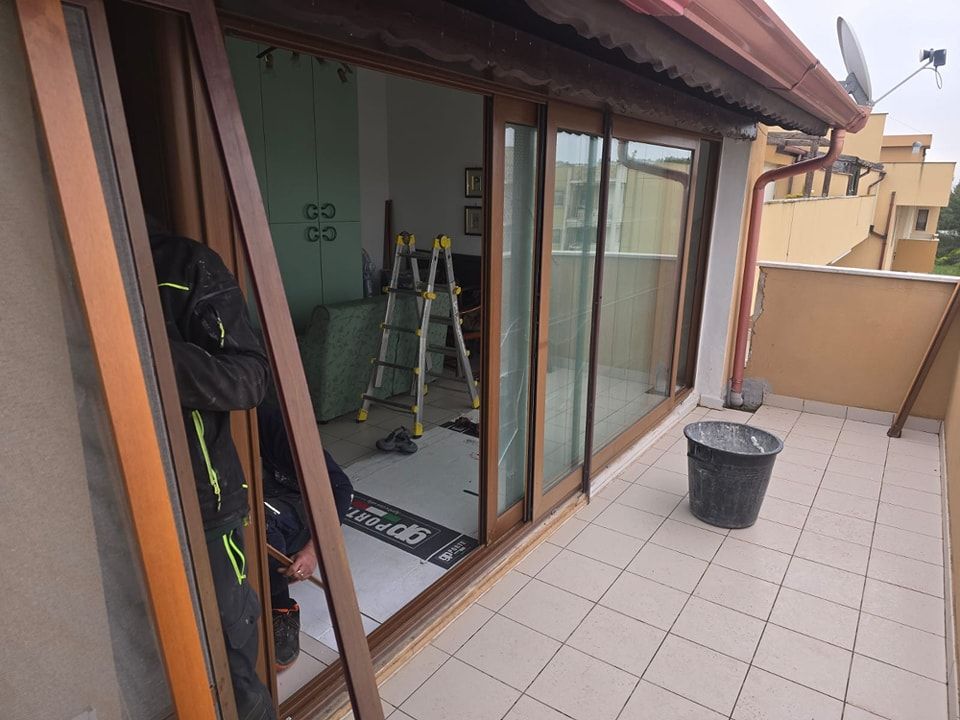 Persona che installa una porta scorrevole in vetro su un balcone; sono visibili cornici marroni, un cestino nero e una scala.