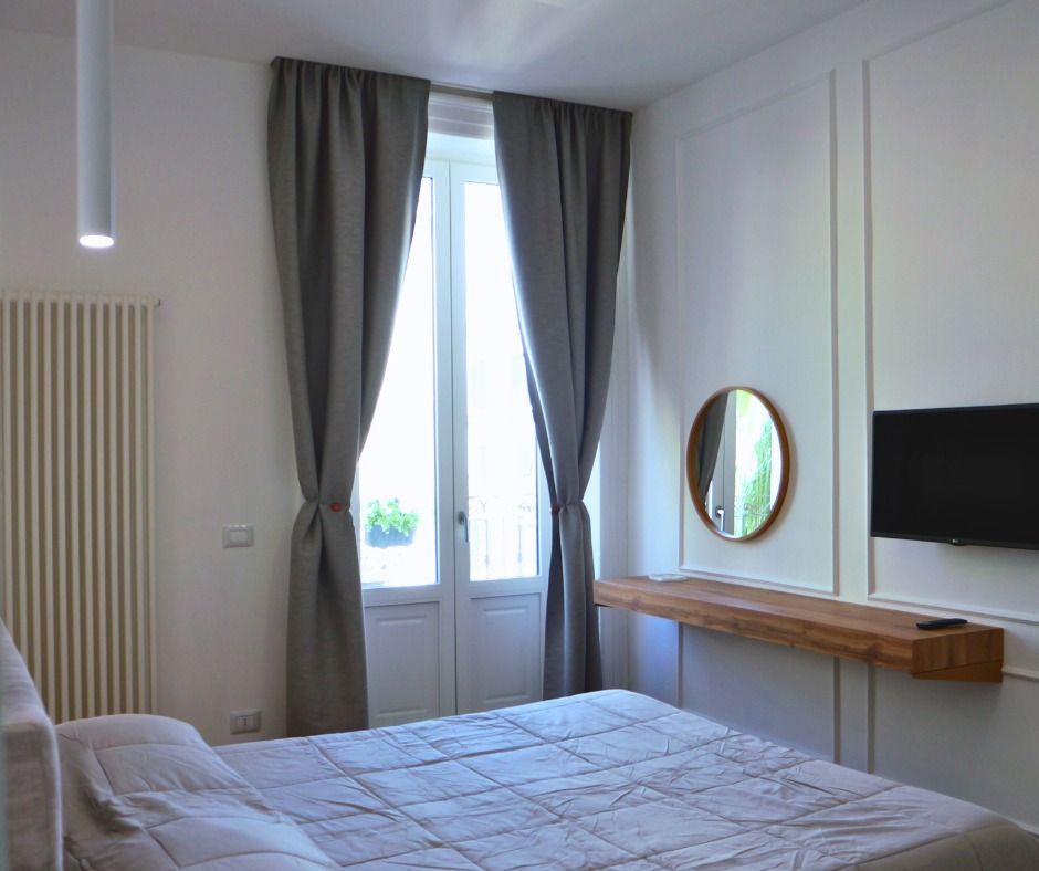 Camera da letto con tende grigie, un letto, una finestra, uno specchio e una TV montata.