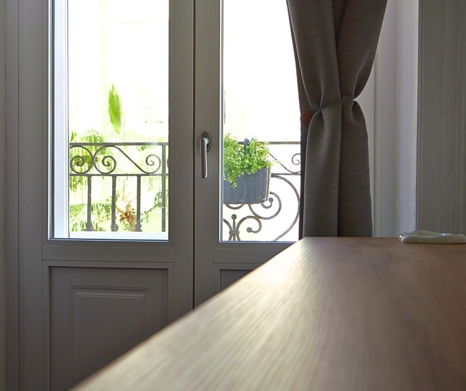 Le porte-finestre si aprono su un balcone con ringhiera decorativa, piante in vaso e luce. Una tenda grigia è tirata.