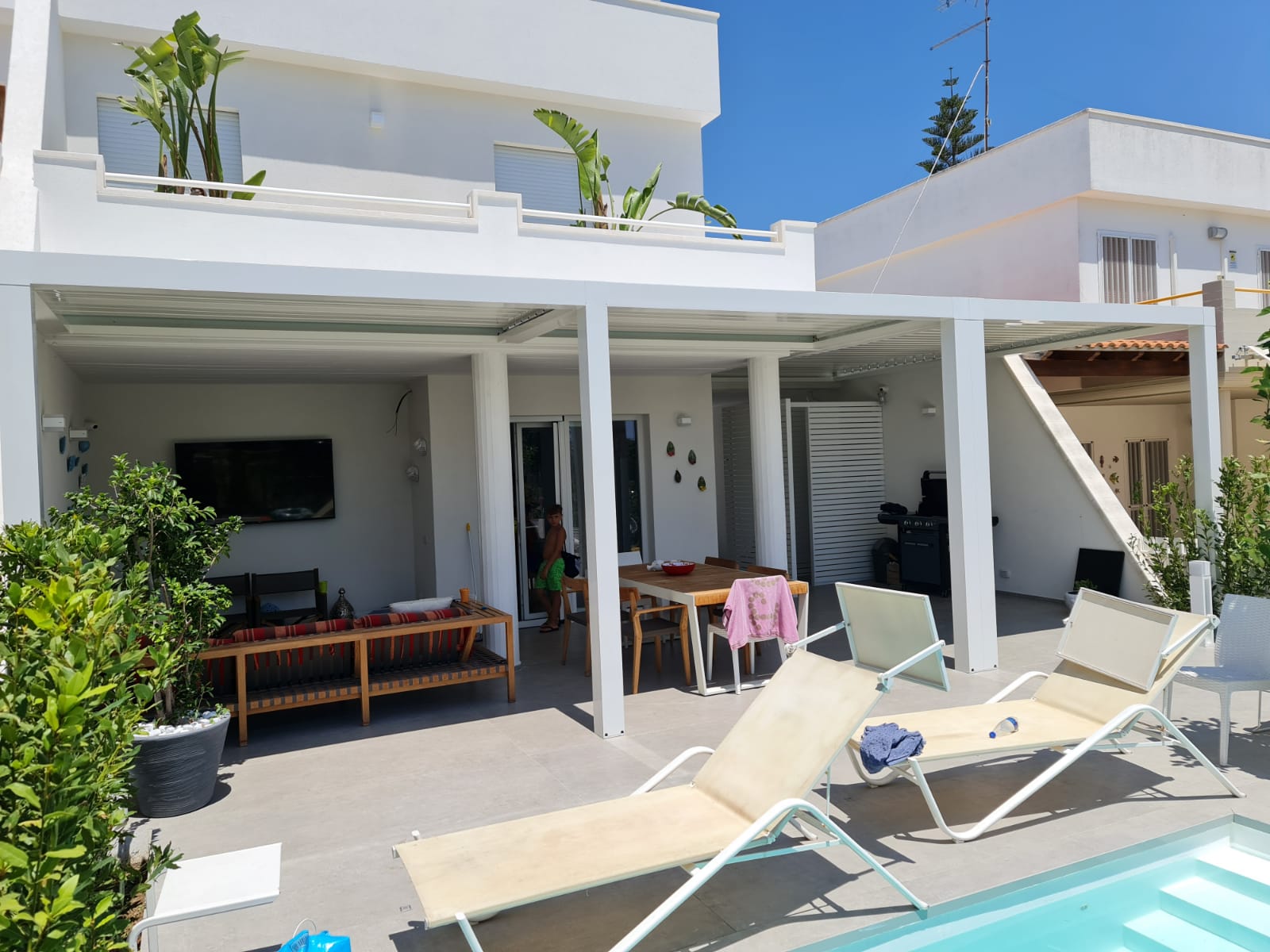 Patio bianco con pergolato, sedie a sdraio, tavolo da pranzo e piscina.