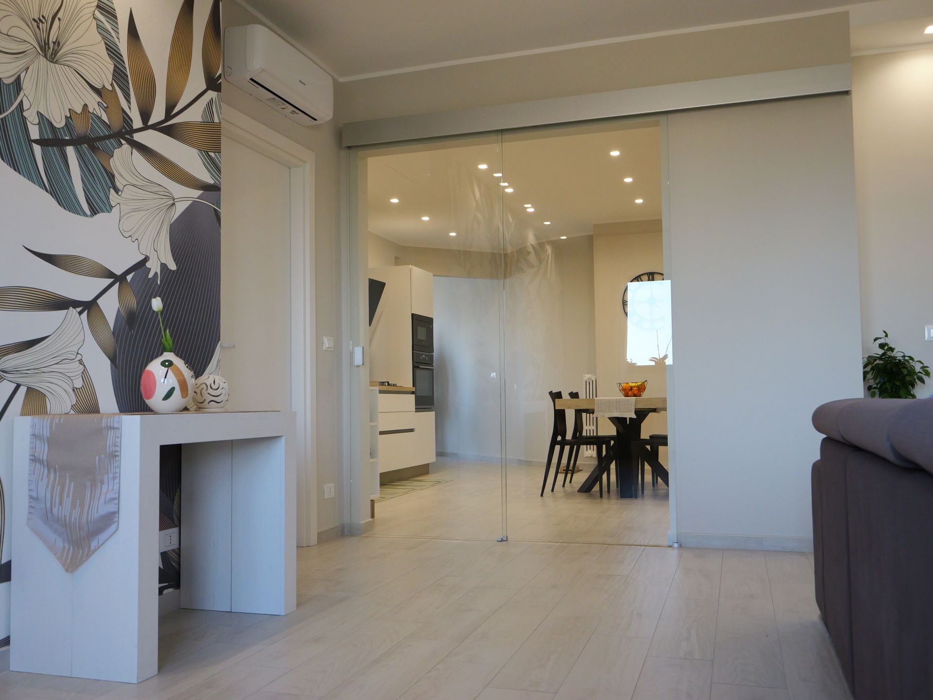 Soggiorno con porta a vetri che conduce alla zona pranzo. Pareti e pavimenti chiari. Consolle decorativa.