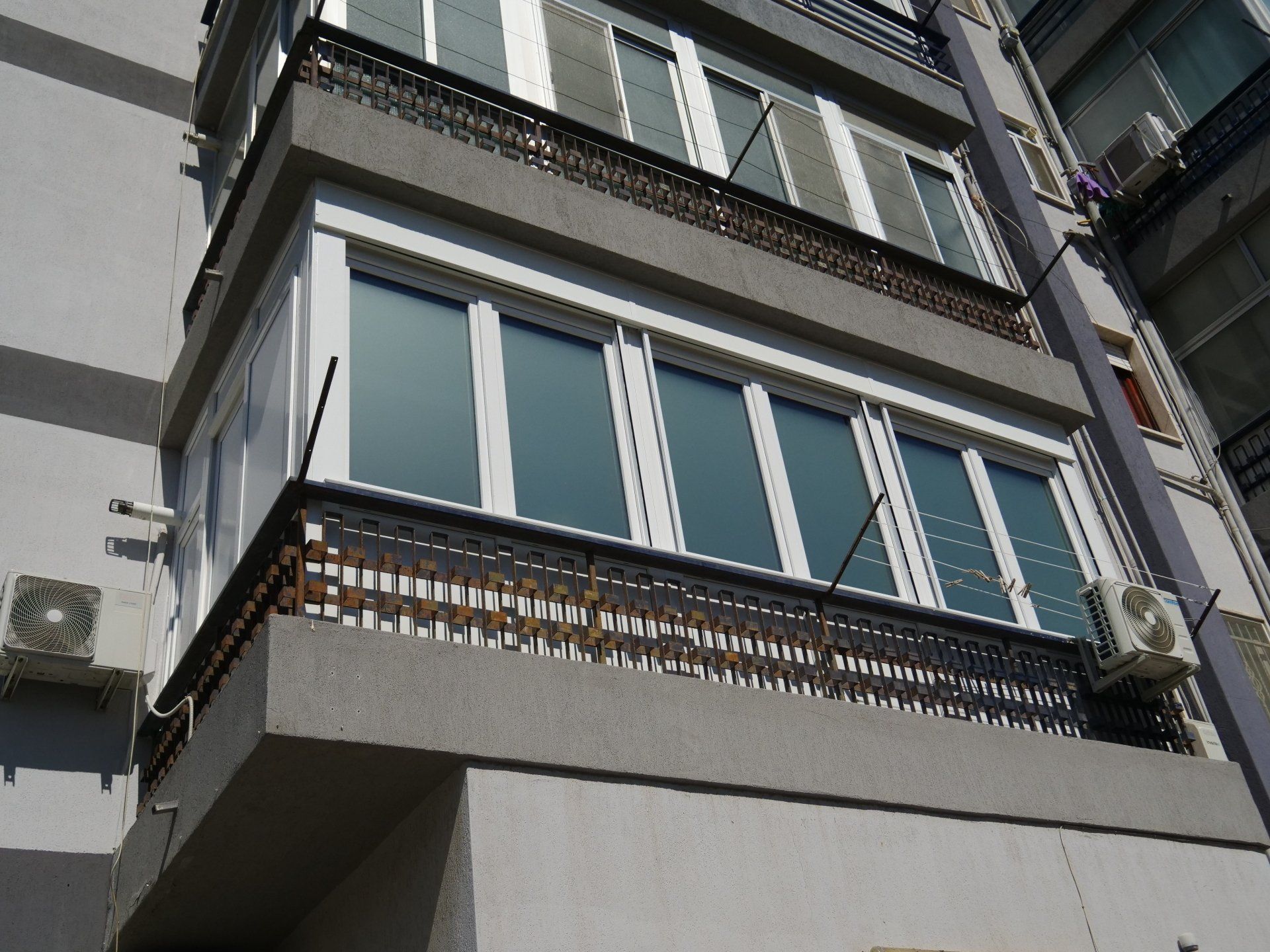 Esterno dell'edificio con balconi. Facciata in cemento grigio, finestre con cornice bianca, ringhiere nere, unità di aria condizionata.