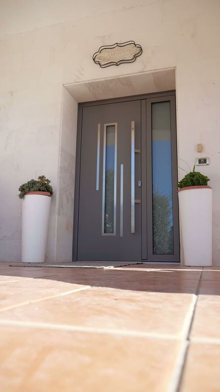 Porta d'ingresso grigia con accenti verticali argentati, fiancheggiata da fioriere bianche.