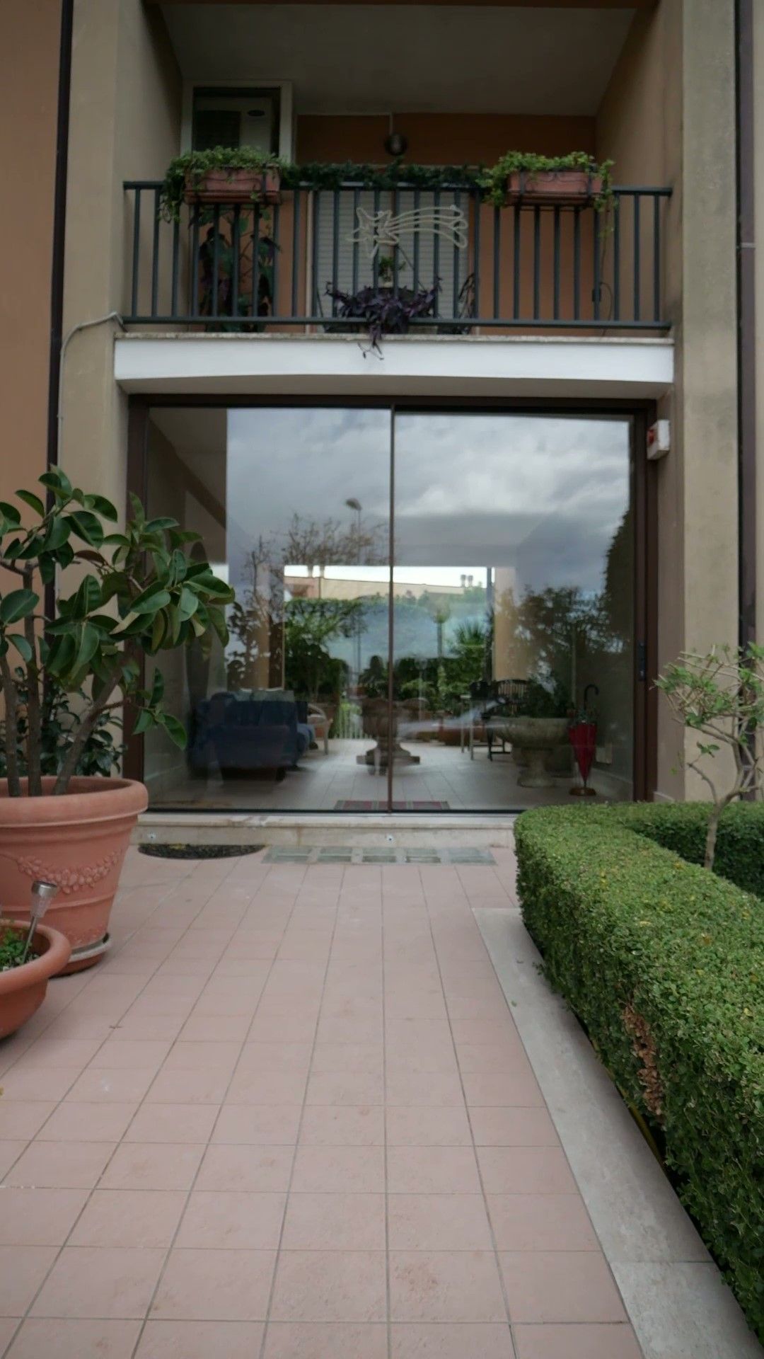 Porte a vetri che conducono a una stanza arredata con mobili e piante su entrambi i lati. Balcone sopra, con piante in vaso.