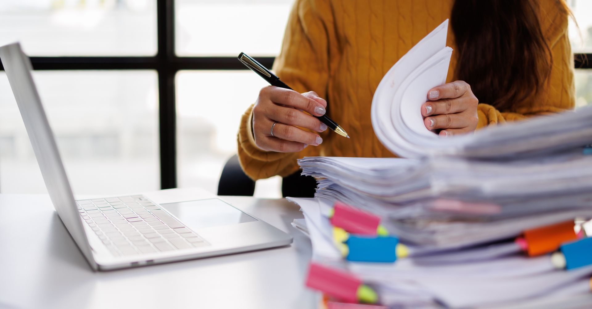 Person writing on papers beside a laptop and a tall stack of documents on a desk