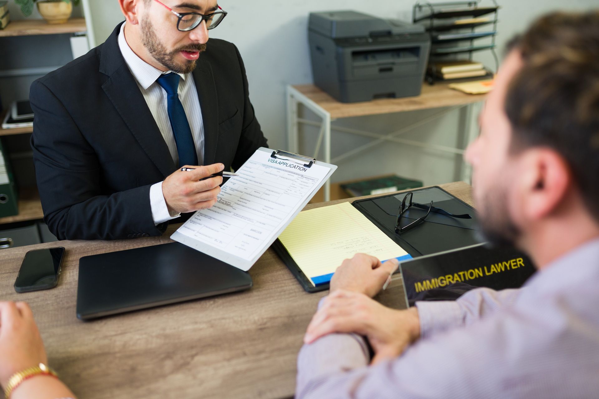 Two men in a business meeting reviewing documents at a desk, with a laptop and office printer nearby.