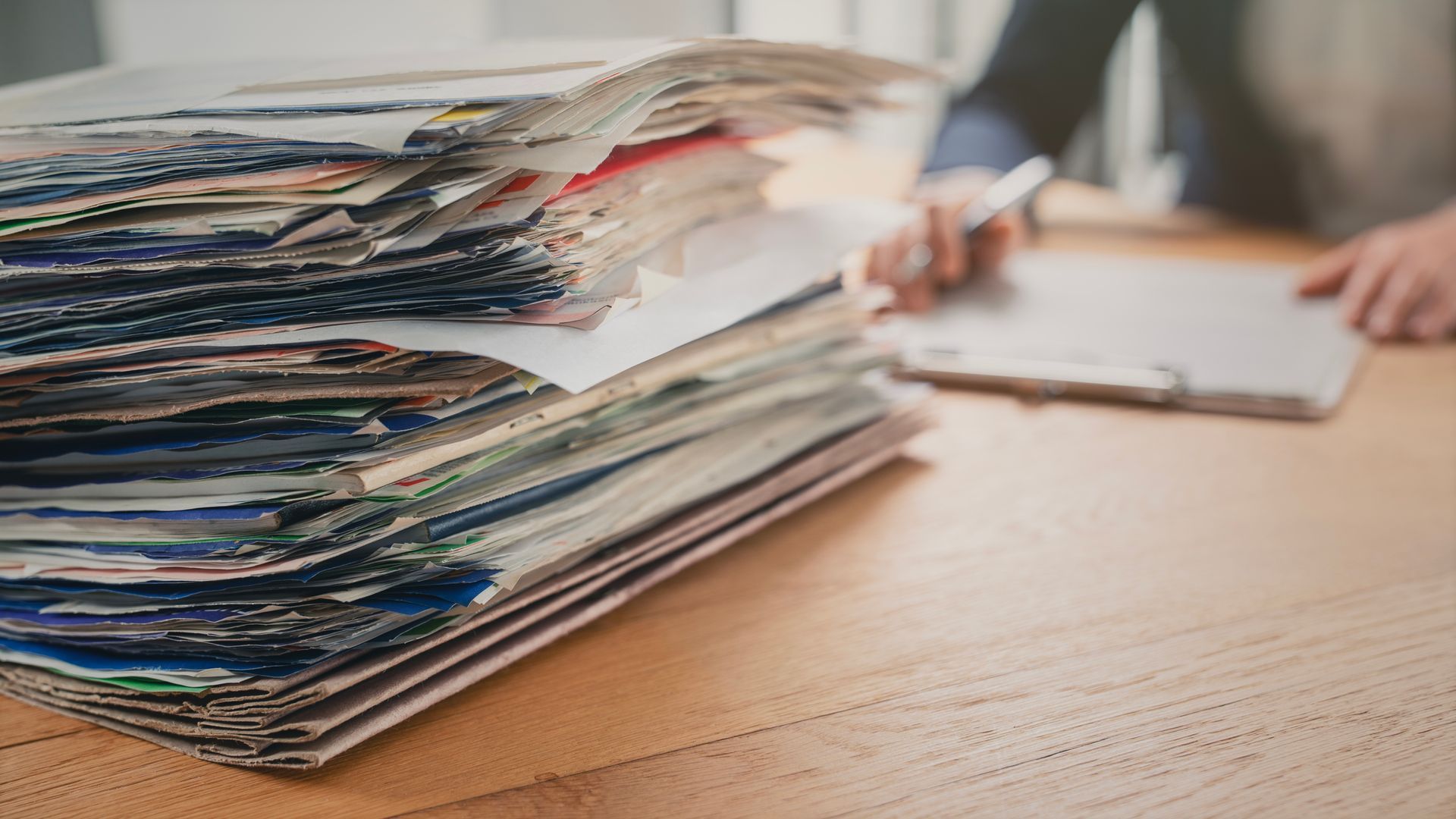Stack of paper files on a wooden desk with a blurred person writing in the background