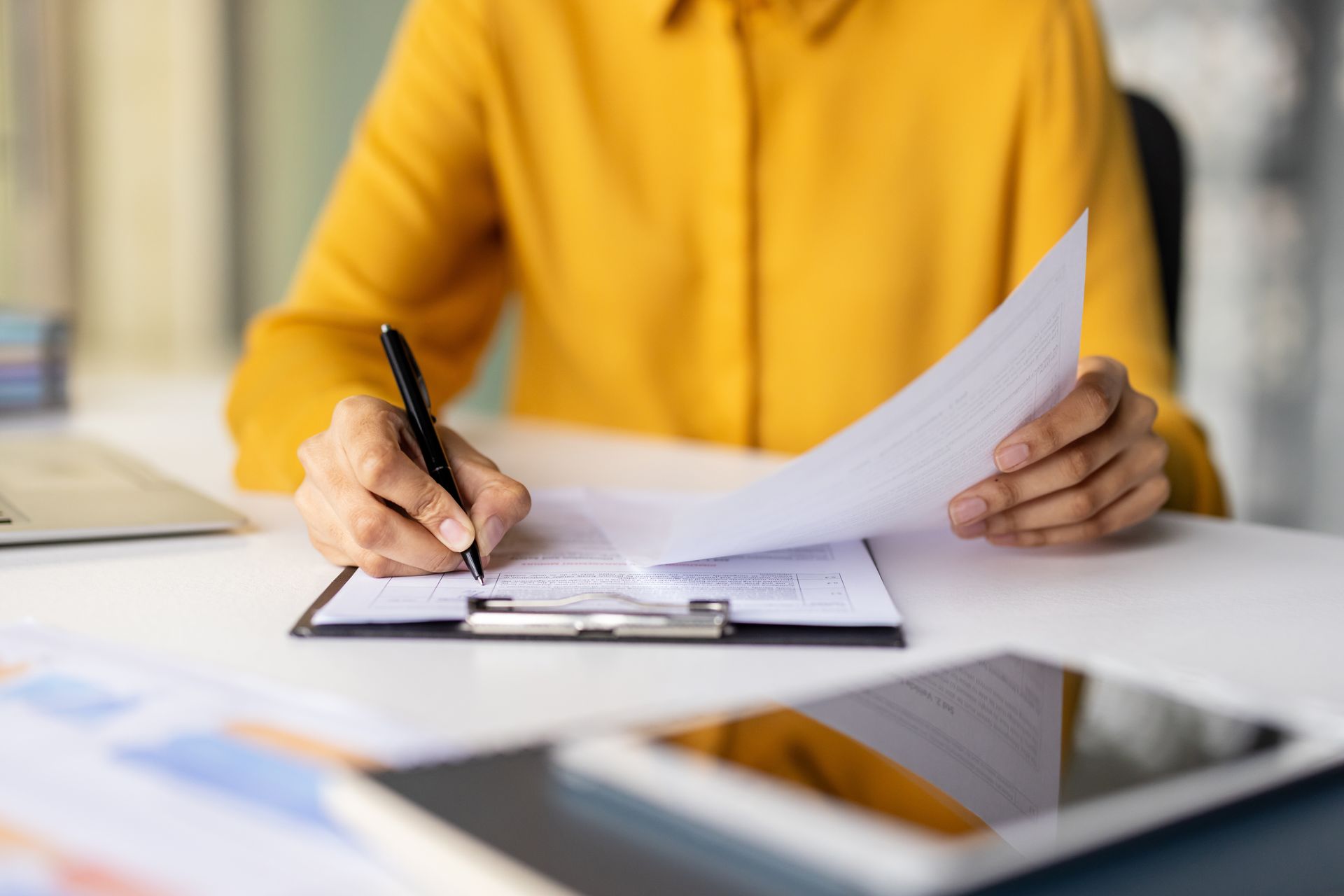 Person in a yellow sweater writing on paper at a desk with a clipboard and tablet nearby