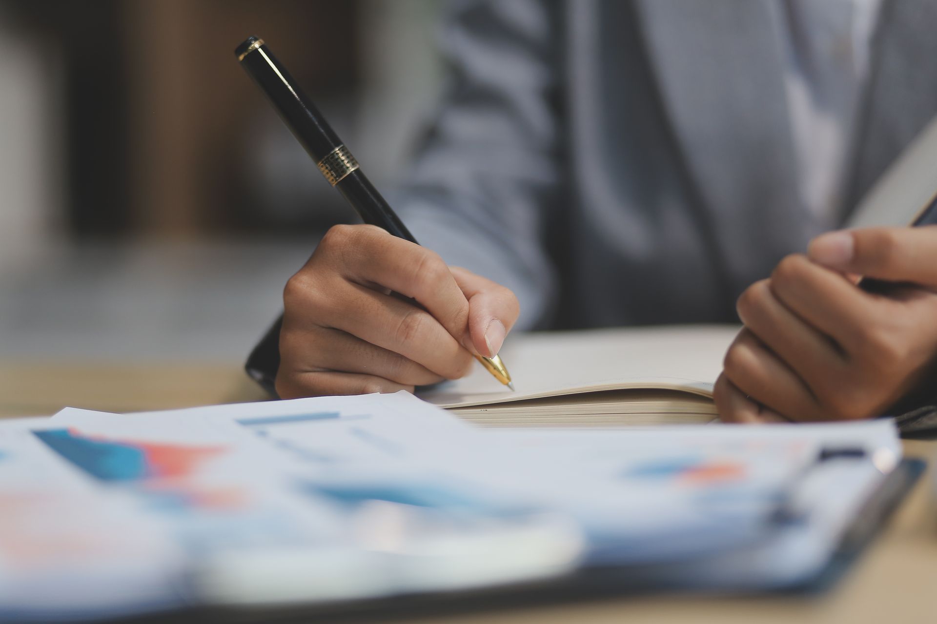Hands writing with a pen on paper at a desk, with documents in the foreground.