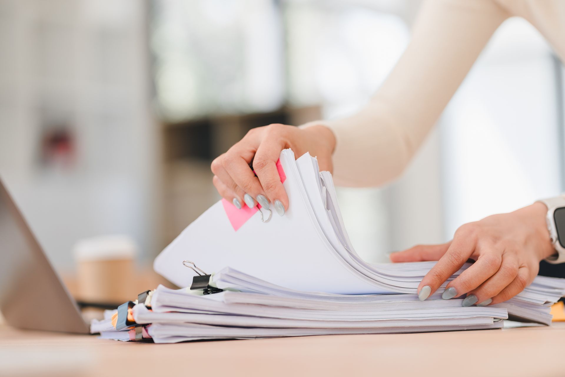 Hands flipping through a stack of papers on a desk beside a laptop.