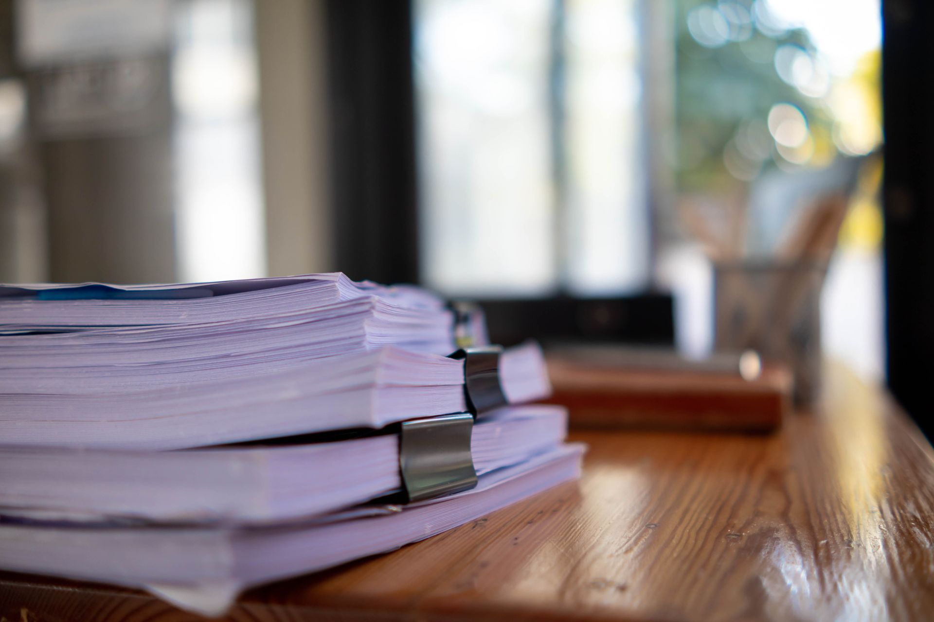 Stack of papers with binder clips on a wooden table near a bright window