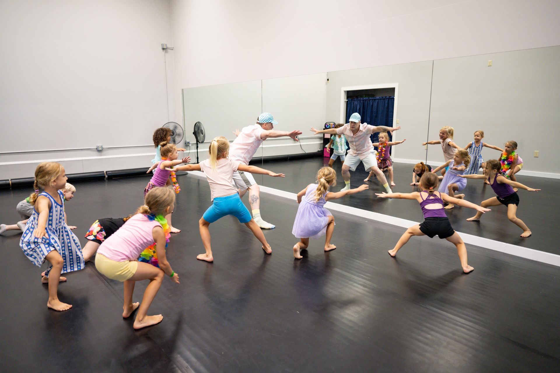 Children practicing a dance routine in a studio, crouched in a wide stance on a black mat.
