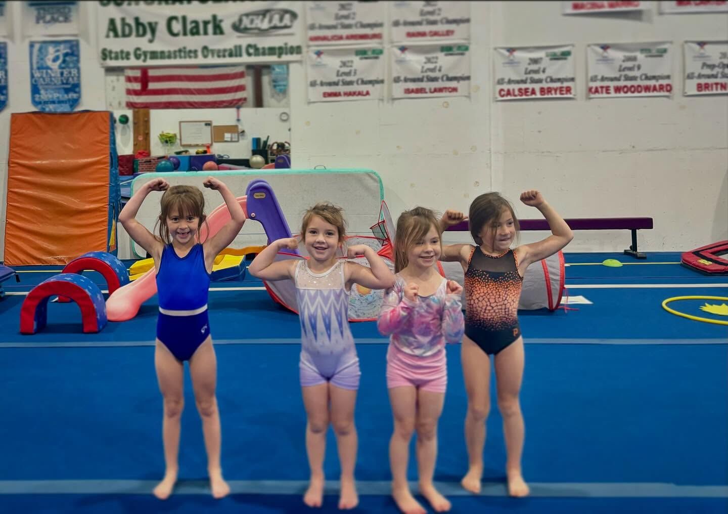 Four young gymnasts posing on a blue mat in a gym, flexing their arms and smiling