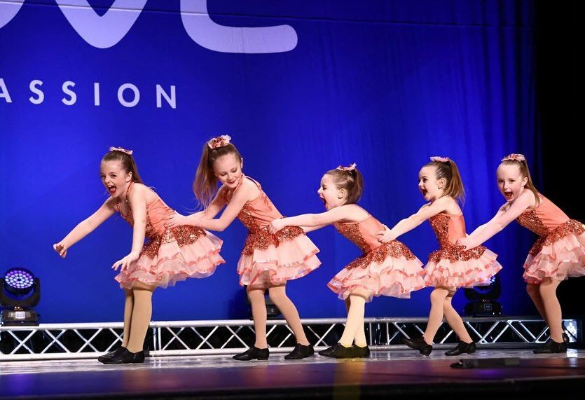 Five young dancers in peach costumes perform on stage under blue lights.