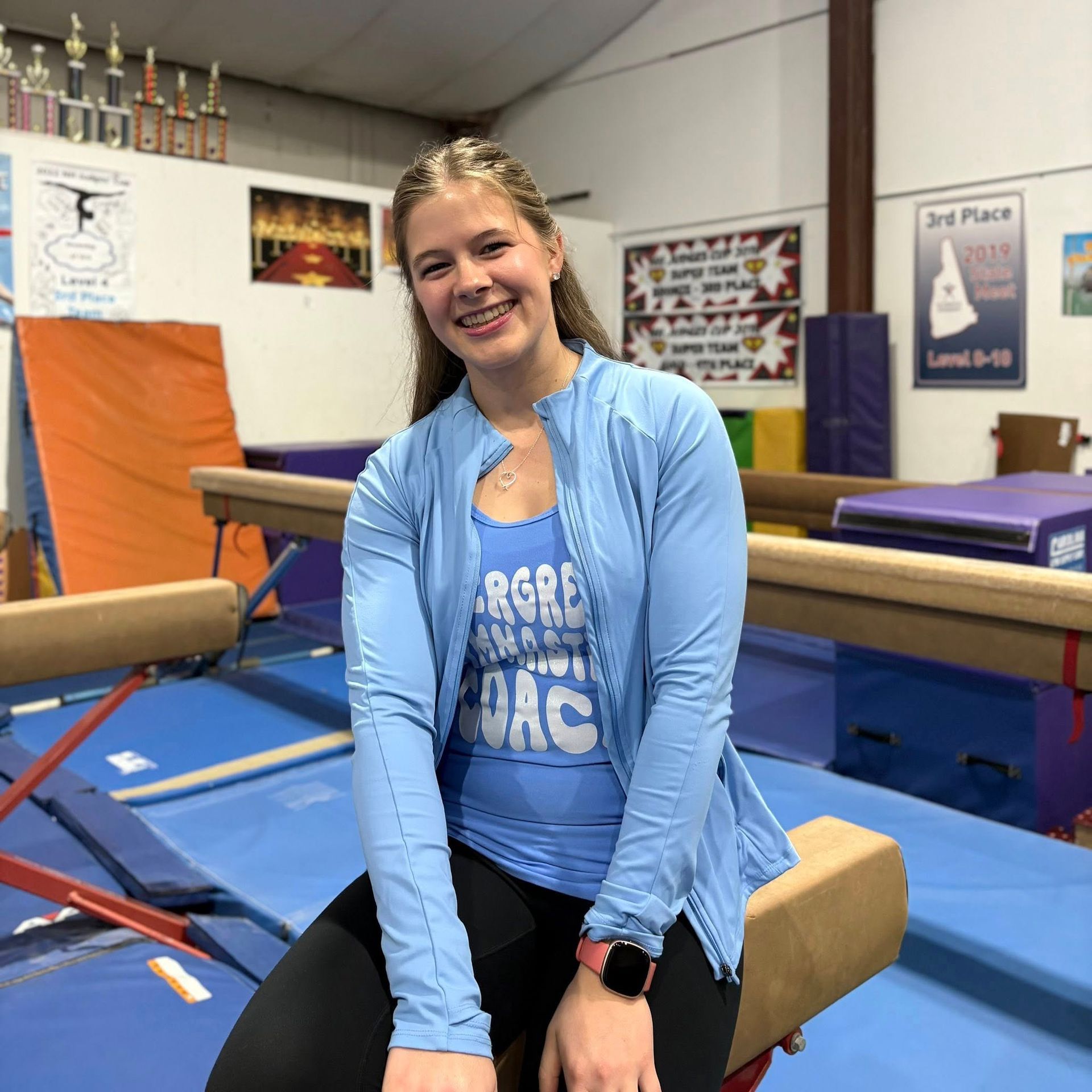 Smiling person sitting on gymnastics equipment in a gym, wearing a light blue top and black leggings.