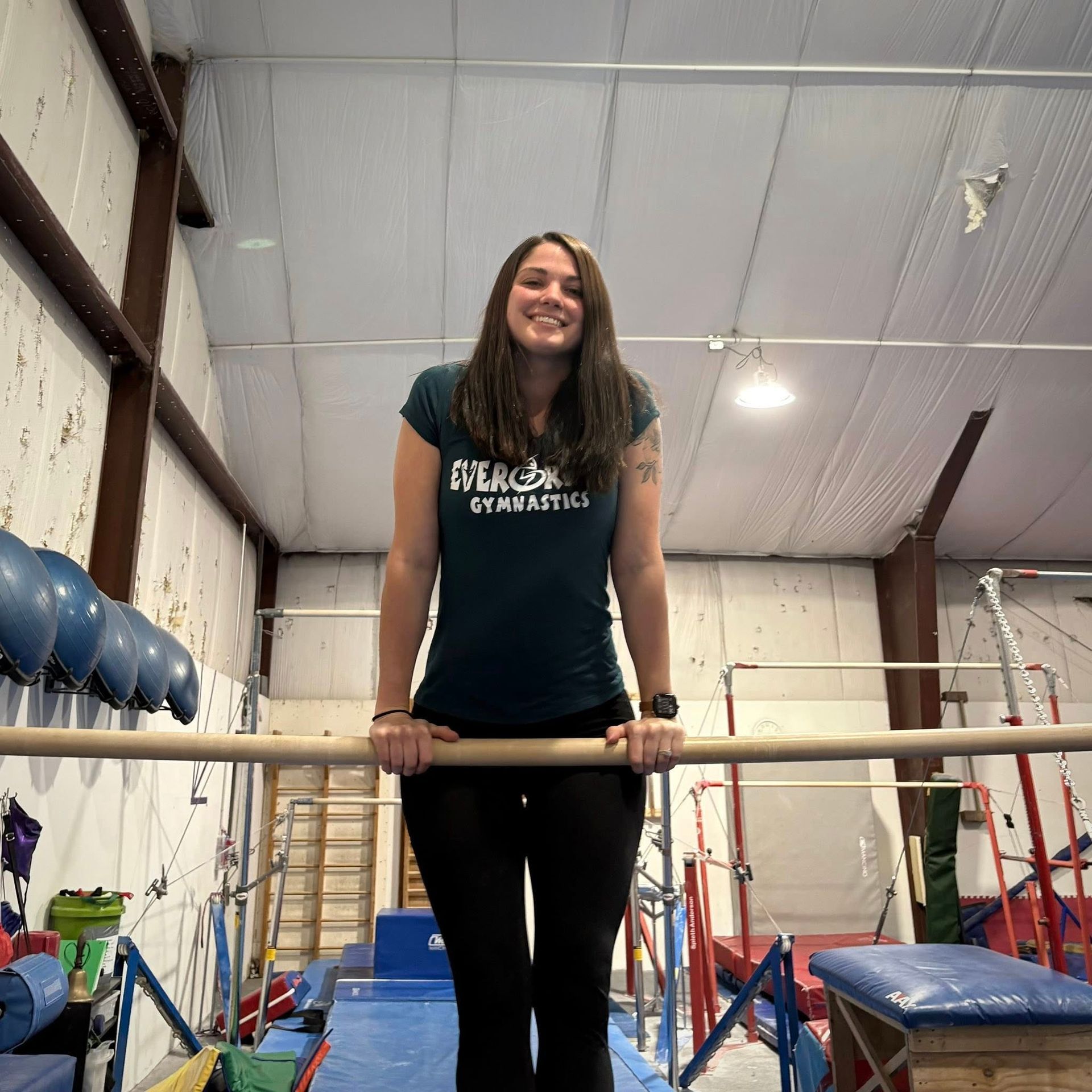 Woman in a gym holding a bar, smiling beside blue mats and equipment.