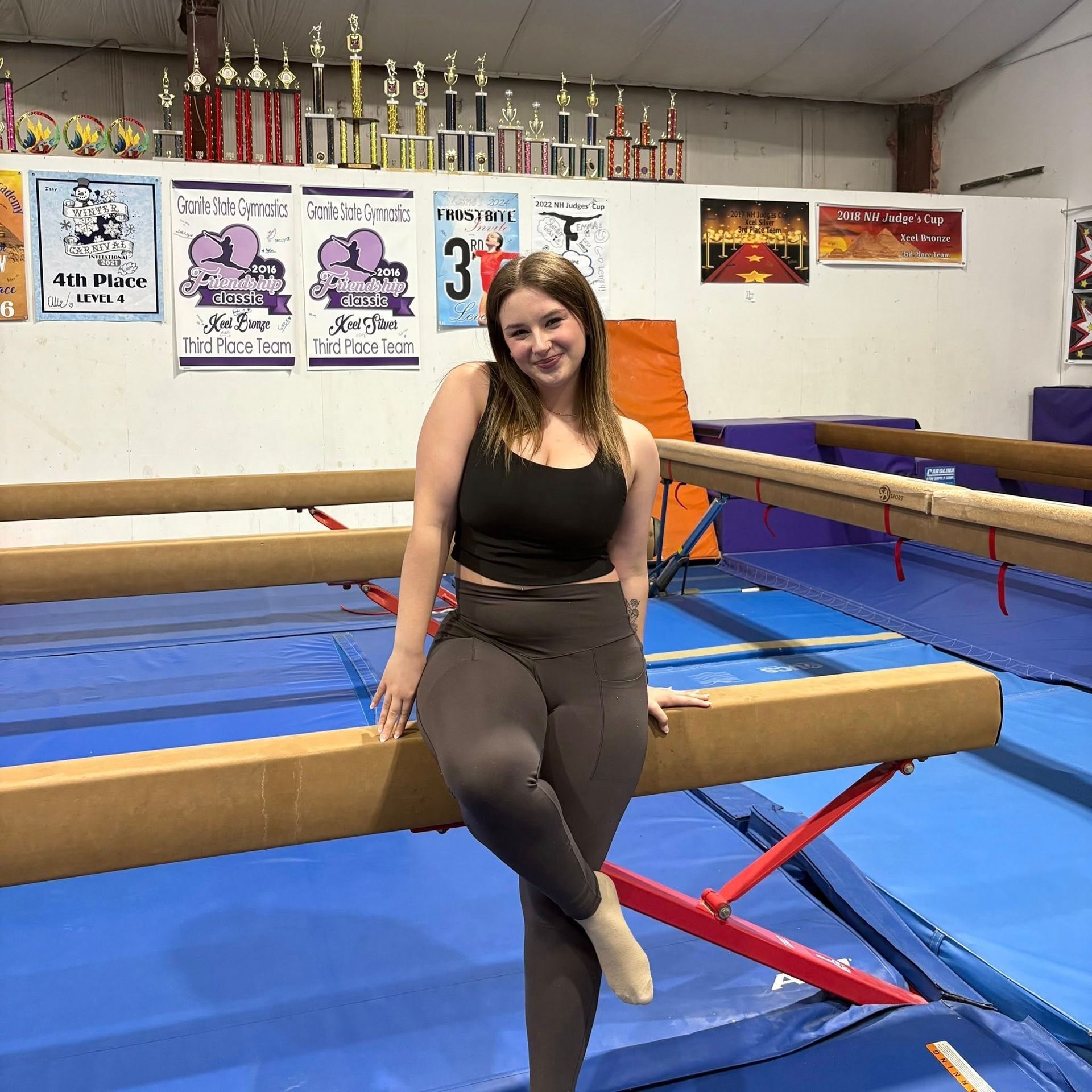 Woman sitting on a boxing ring rope in a gym, with blue mats and trophies in the background