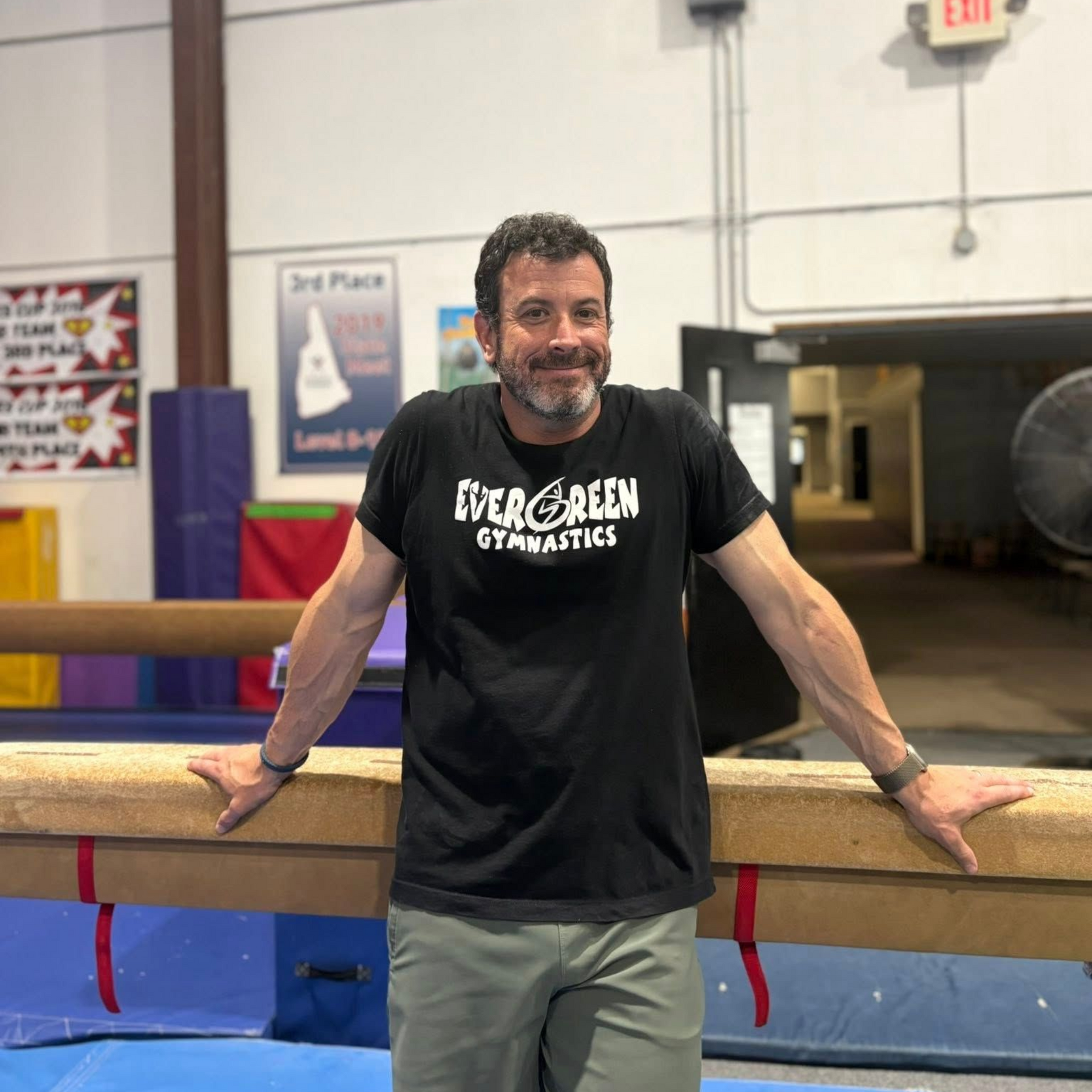 Man in black sleeveless shirt leaning on a gym balance beam, smiling in an indoor gymnastics facility.