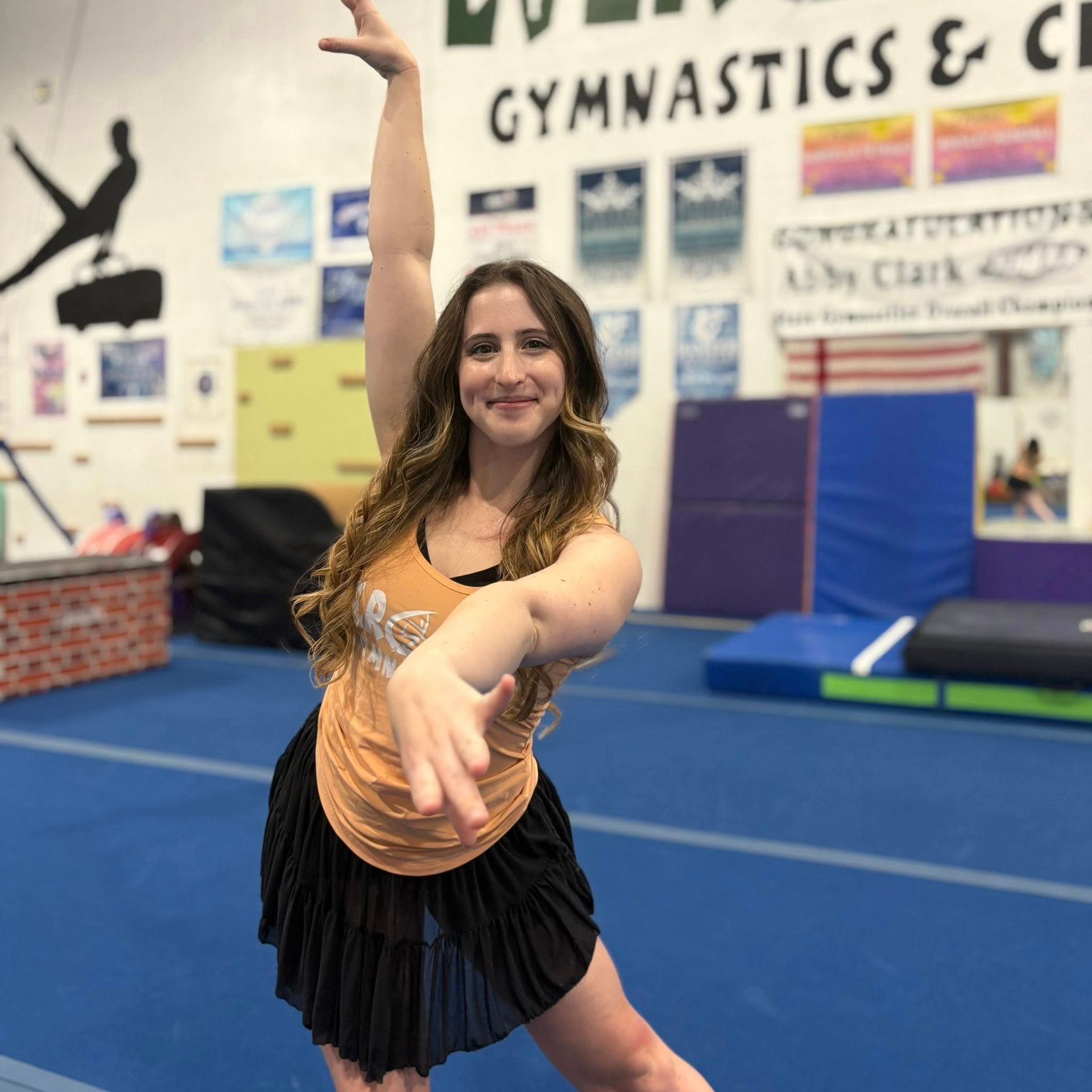 Gymnast posing on blue mat in a gymnastics gym, smiling with one arm raised and one arm extended.