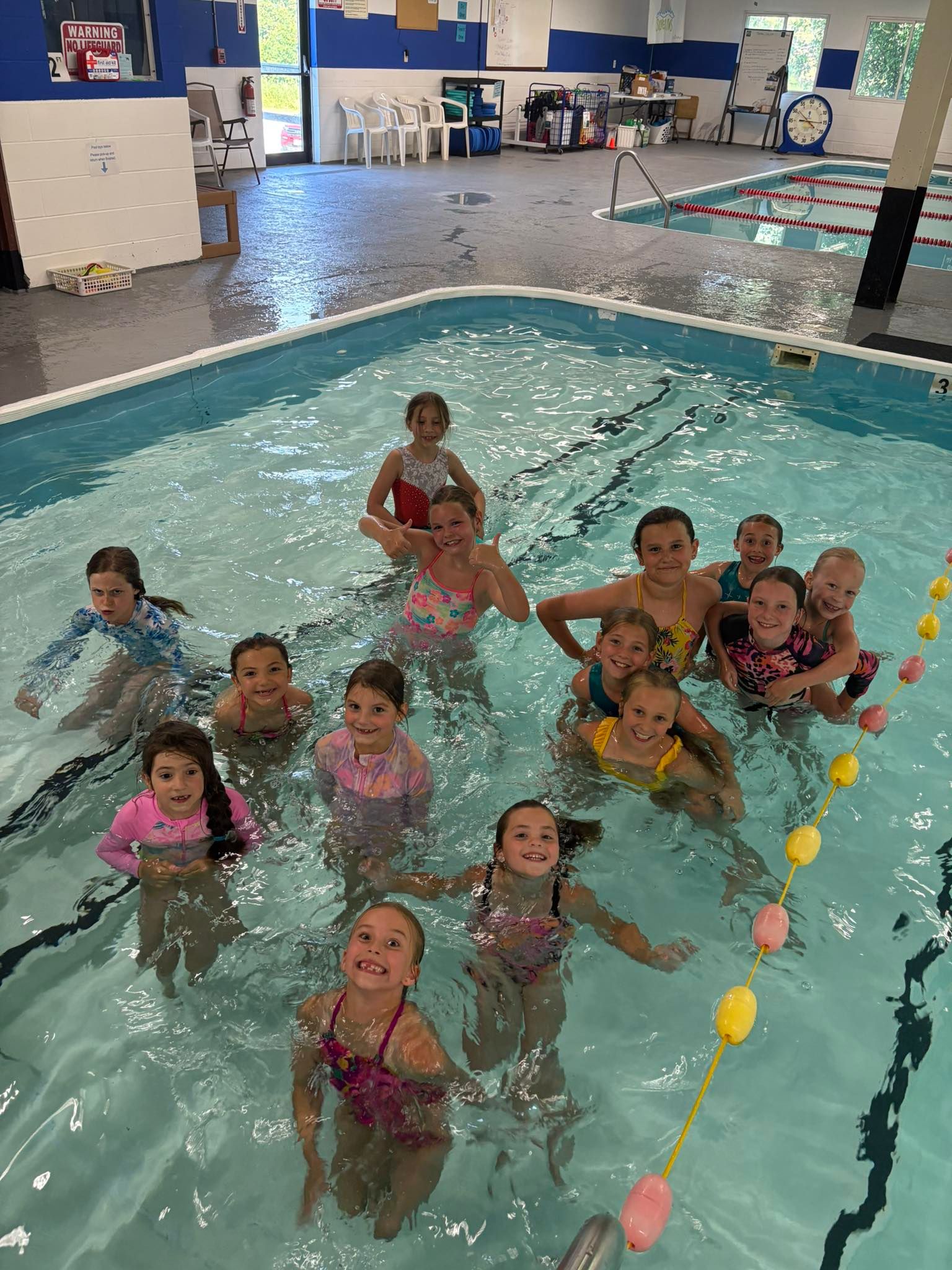 Children playing in an indoor pool with colorful floaties and lane markers, smiling near the water’s edge
