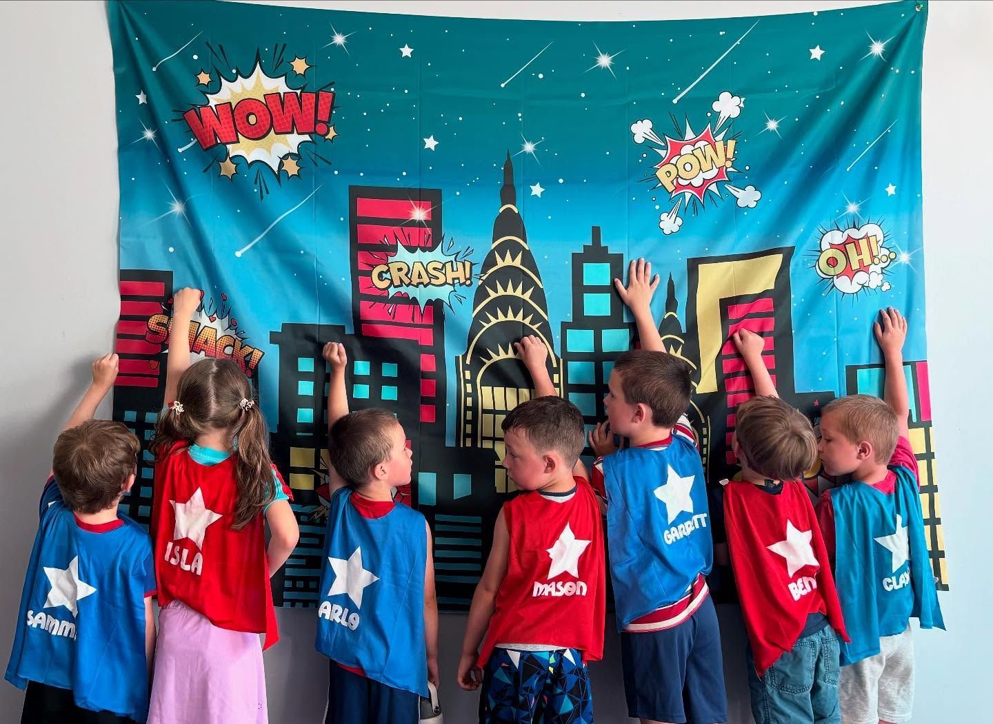 Children in blue and red capes face a comic-style city backdrop, reaching up at a classroom display