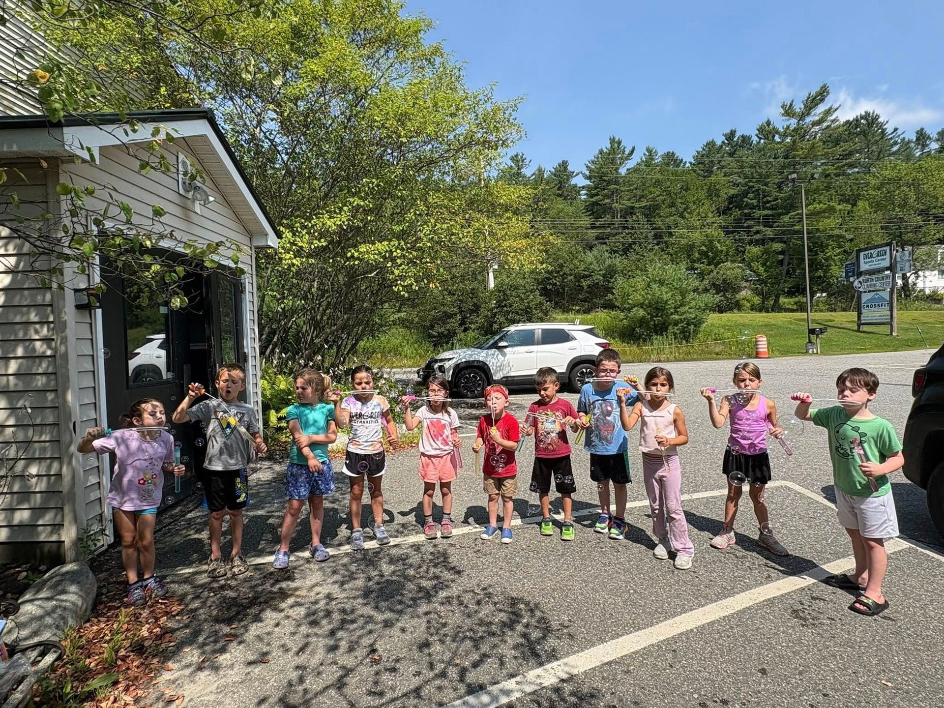 Children lined up outdoors by a building, posing near a road on a sunny day.