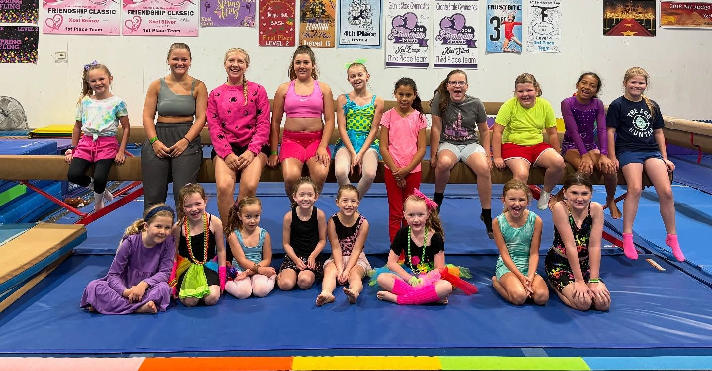 Group of gymnasts posing on mats in a gym, with colorful posters and equipment in the background