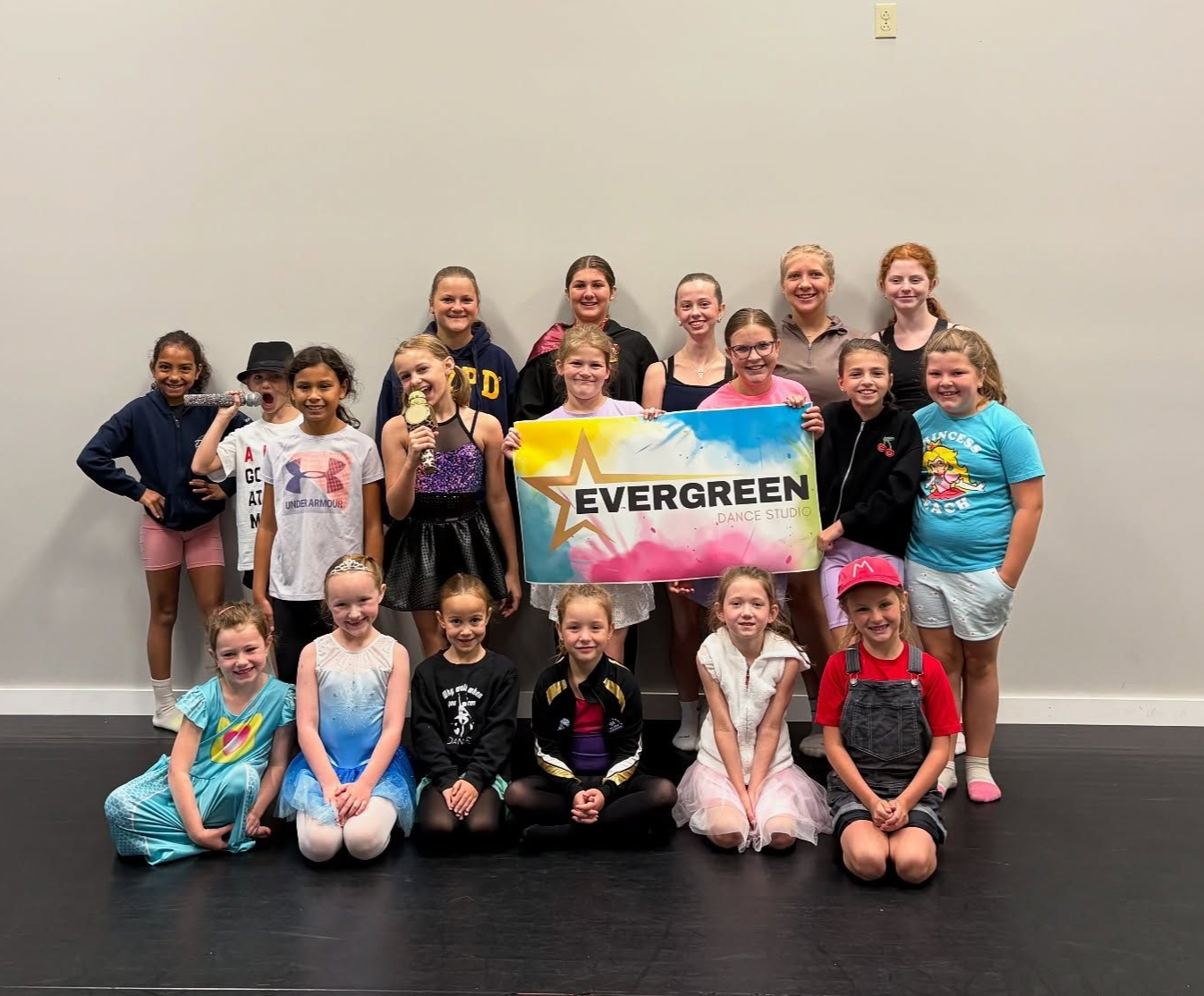 Group of children holding an Evergreen banner in a dance studio, posing together for a team photo.