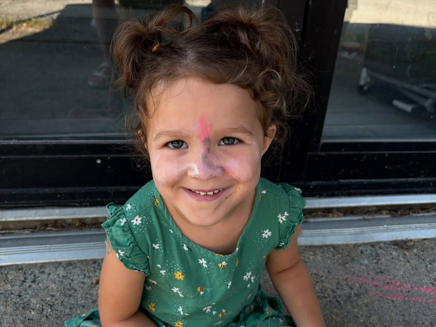 Smiling child with face paint, wearing a green shirt, sitting on steps outdoors near a window