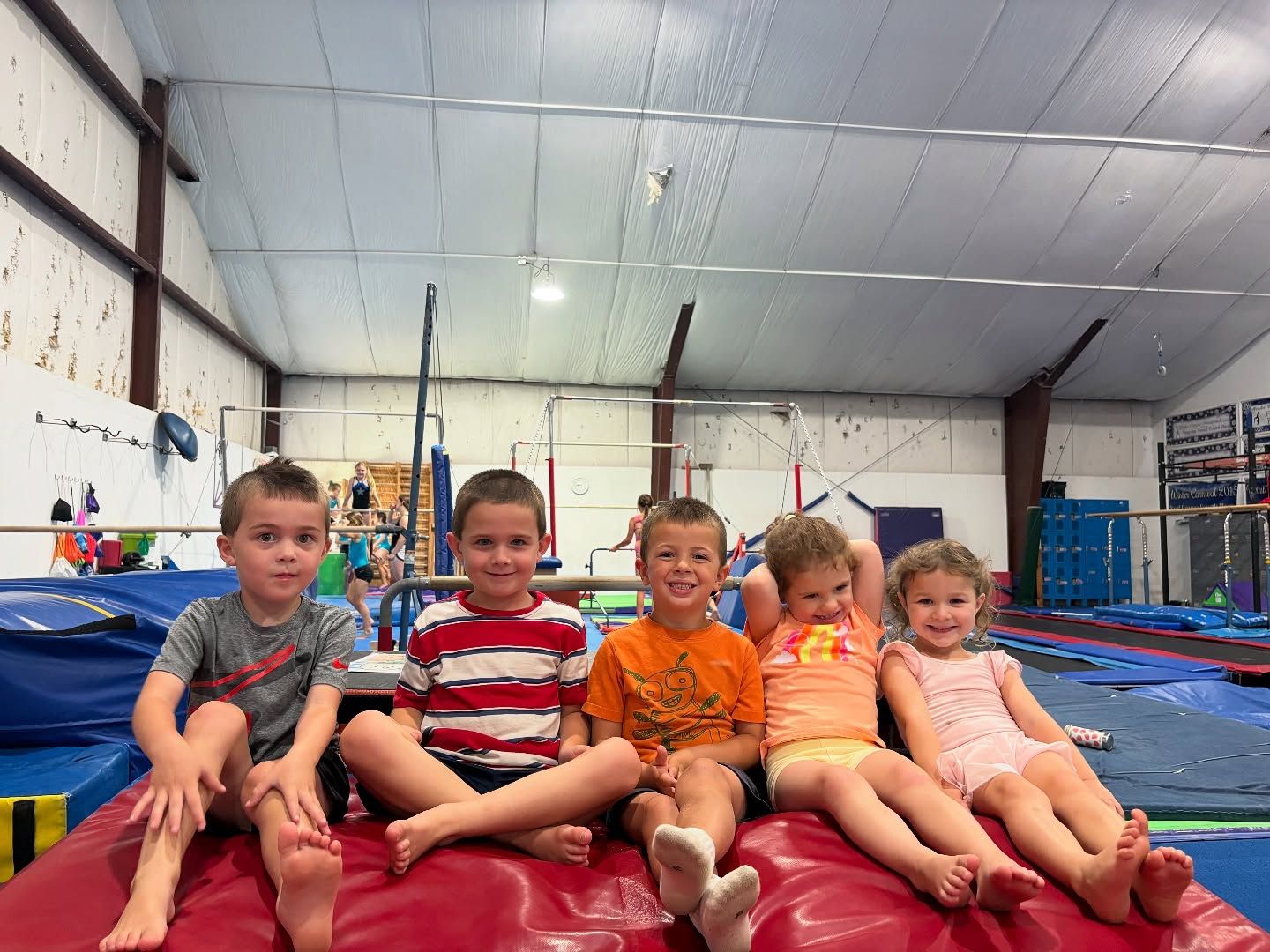 Five children sitting on a red mat inside a gymnastics gym, smiling at the camera.