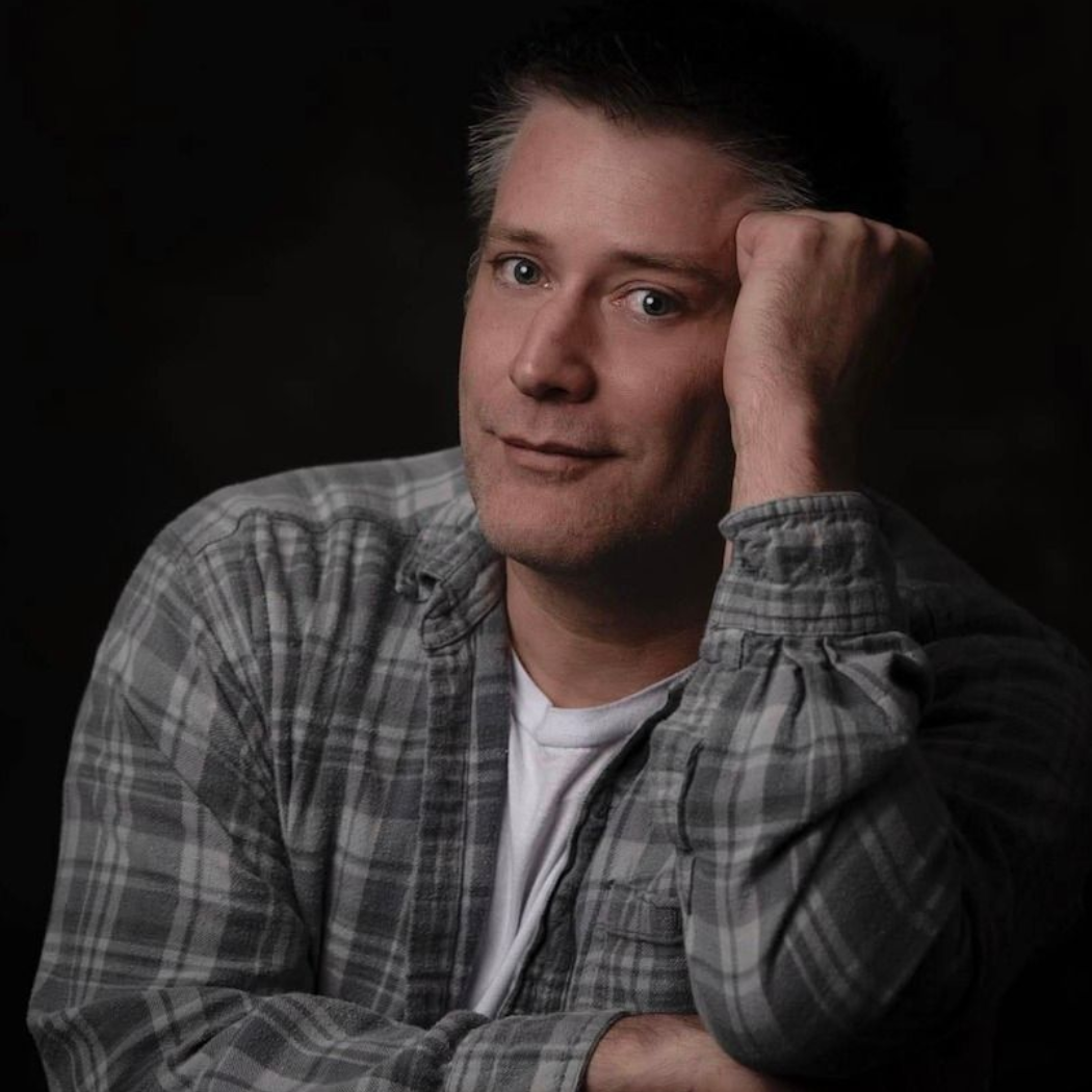 Man in plaid shirt leaning on his hand against a dark background