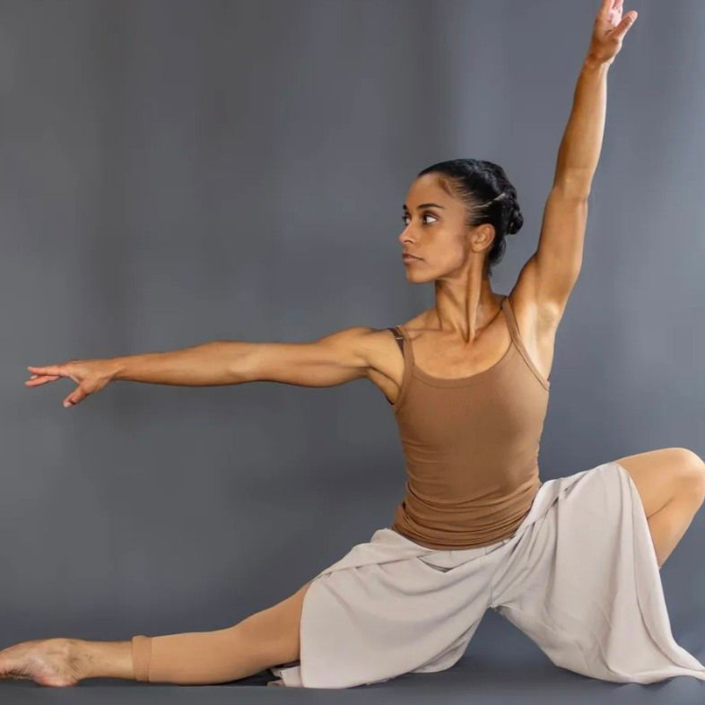 Woman in tan top and beige pants posing in a yoga split against a gray backdrop
