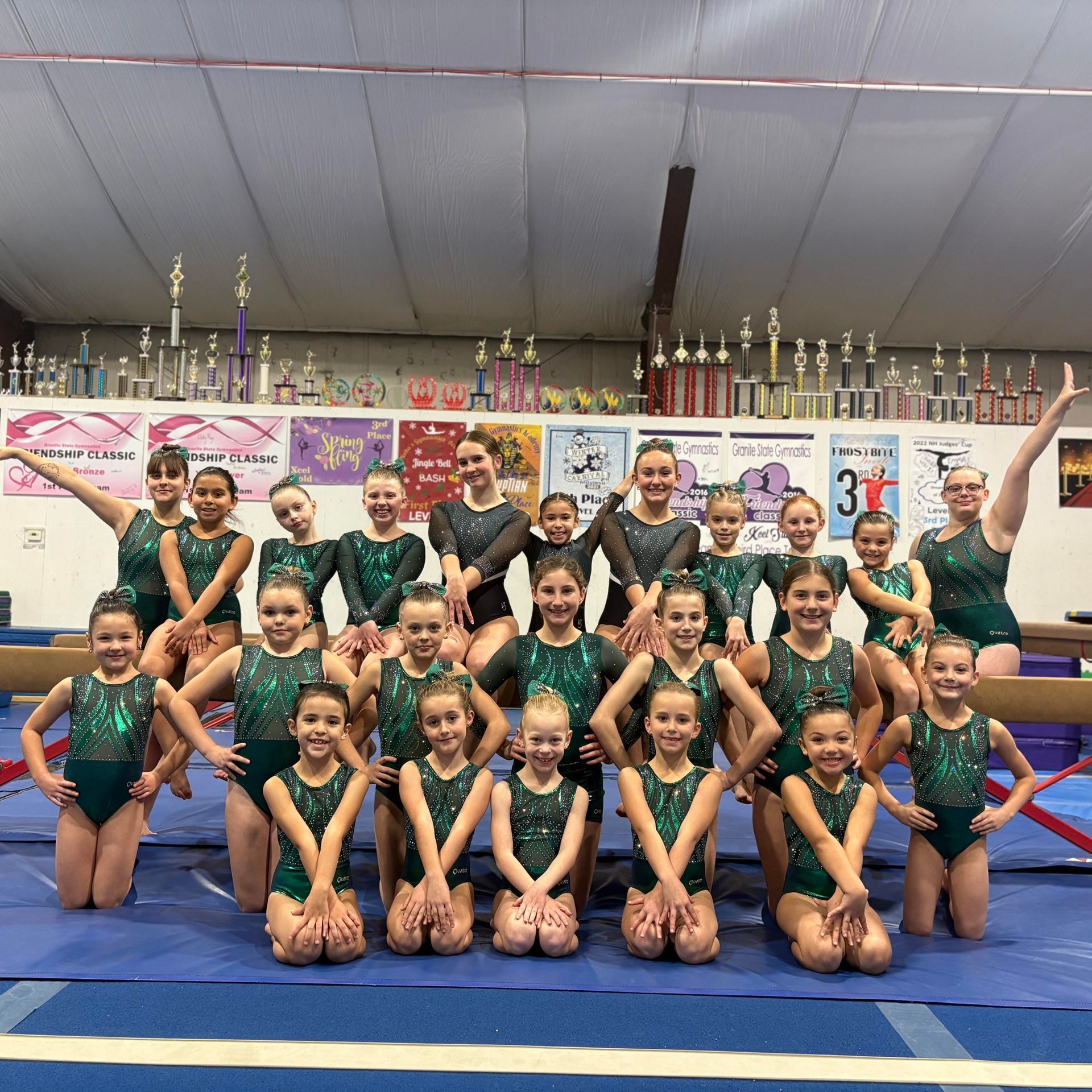 Gymnastics team posing in green leotards on blue mats in a gym, with trophies displayed behind them.