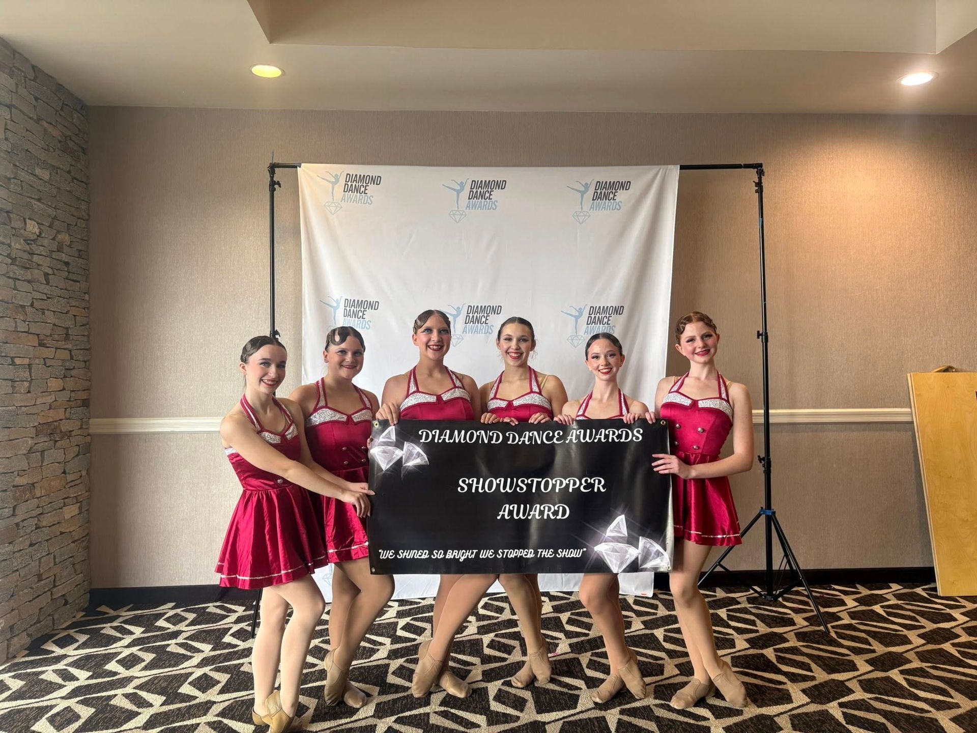 Five dancers in red outfits hold a black sign at an indoor event backdrop.