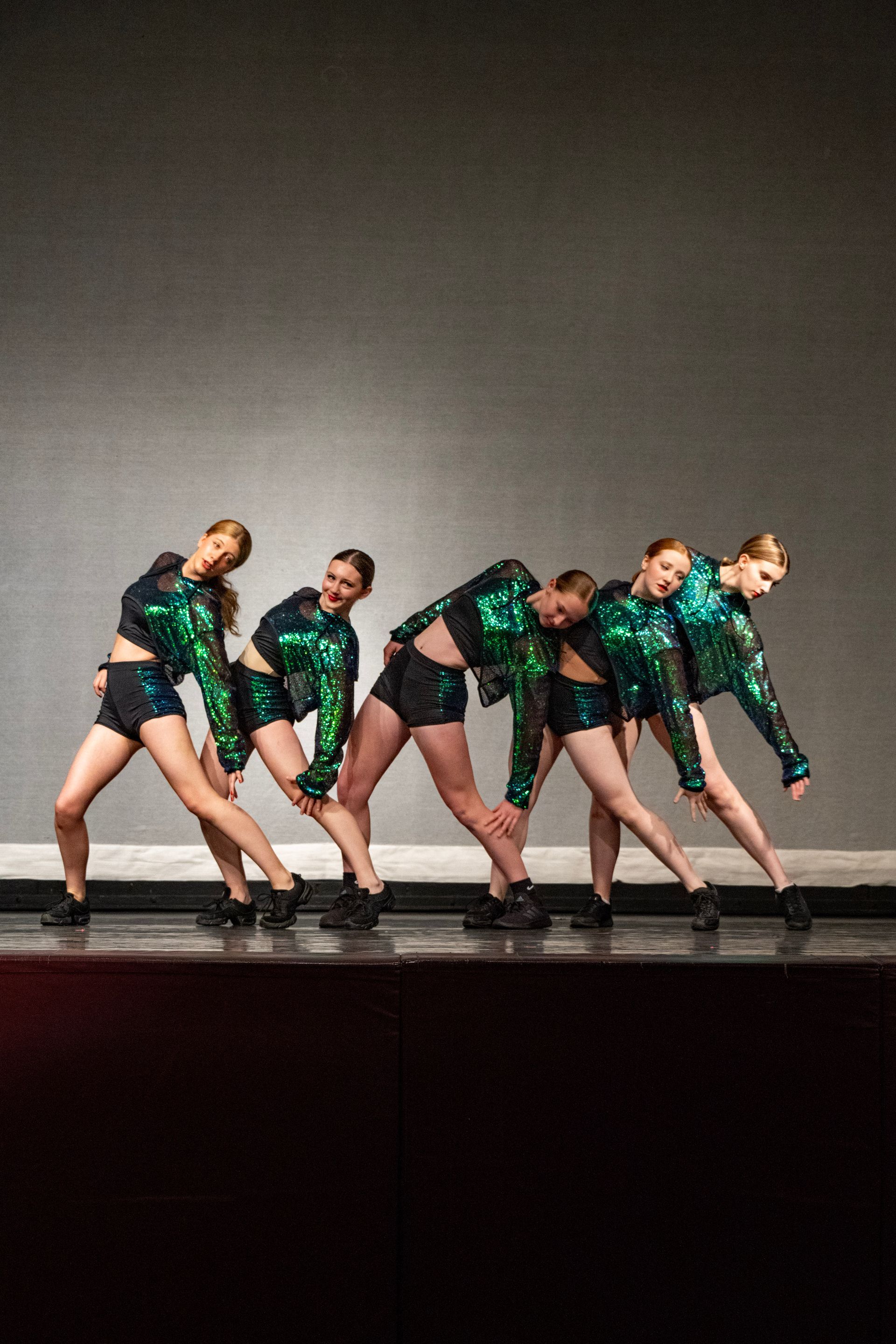 Five dancers in green sequined outfits leaning in a synchronized pose on a stage