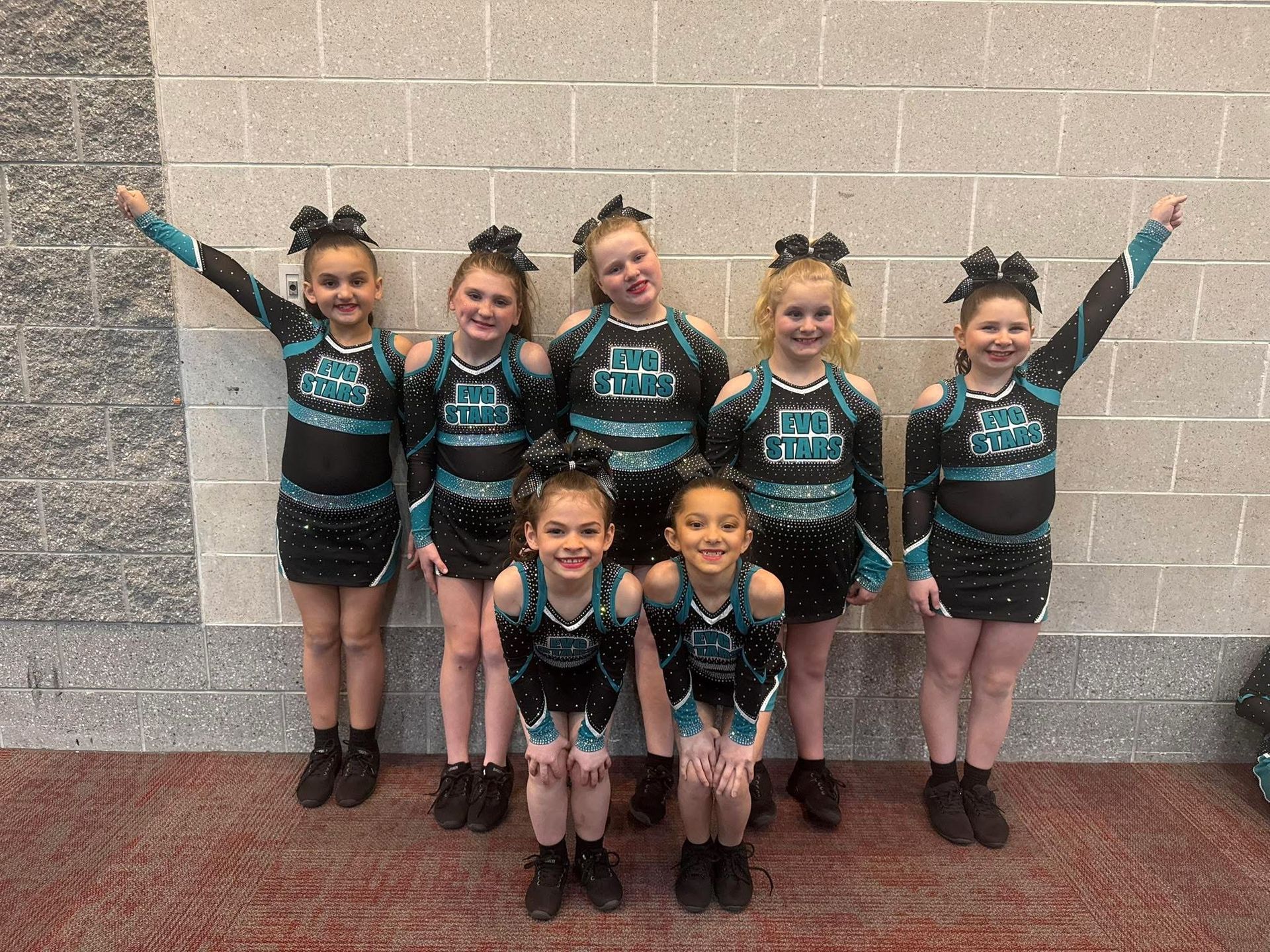 Six children in matching teal and black dance outfits pose against a cinderblock wall