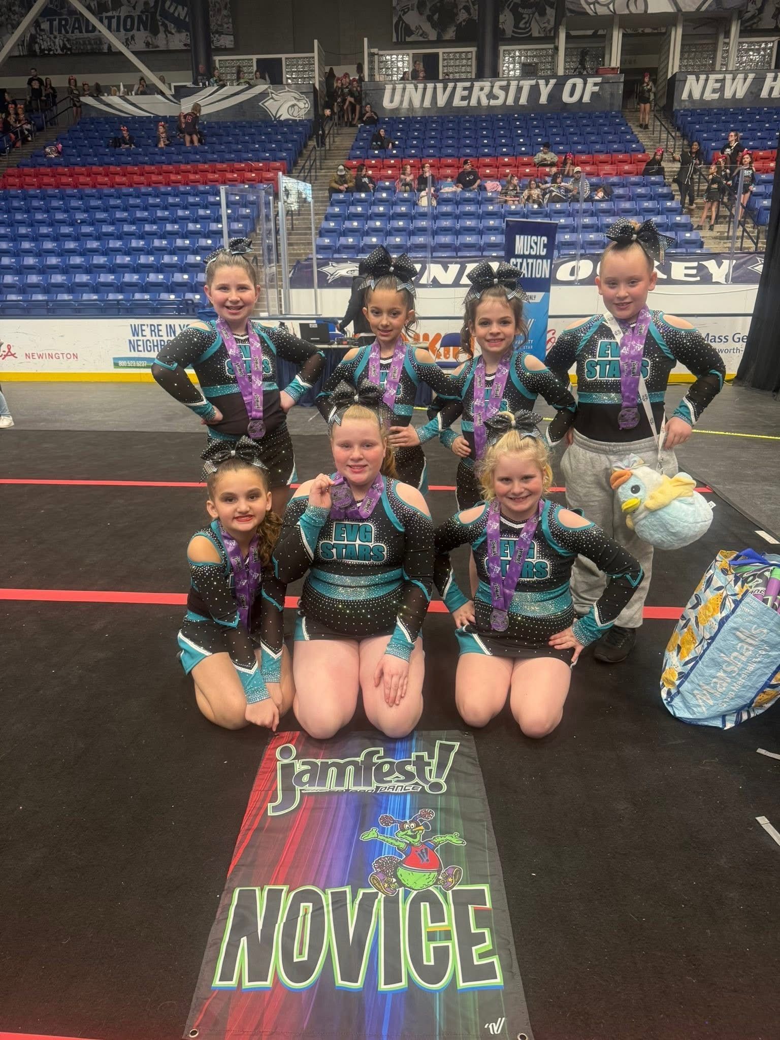 Six cheerleaders in black and teal uniforms pose on a mat that says “NOVICE” in an indoor arena.
