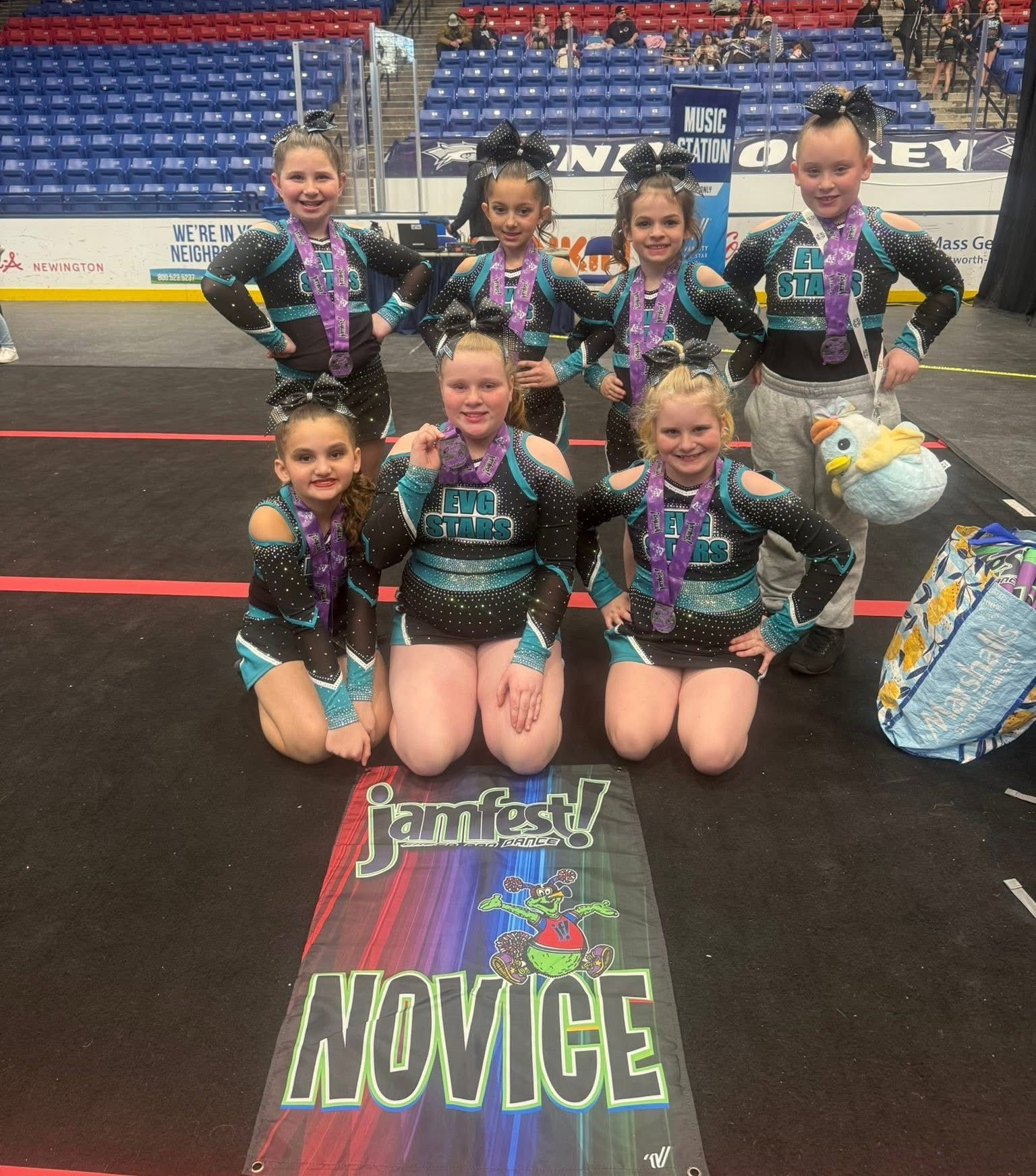 Cheer team posing with a Novice mat in an indoor arena, wearing black and teal uniforms