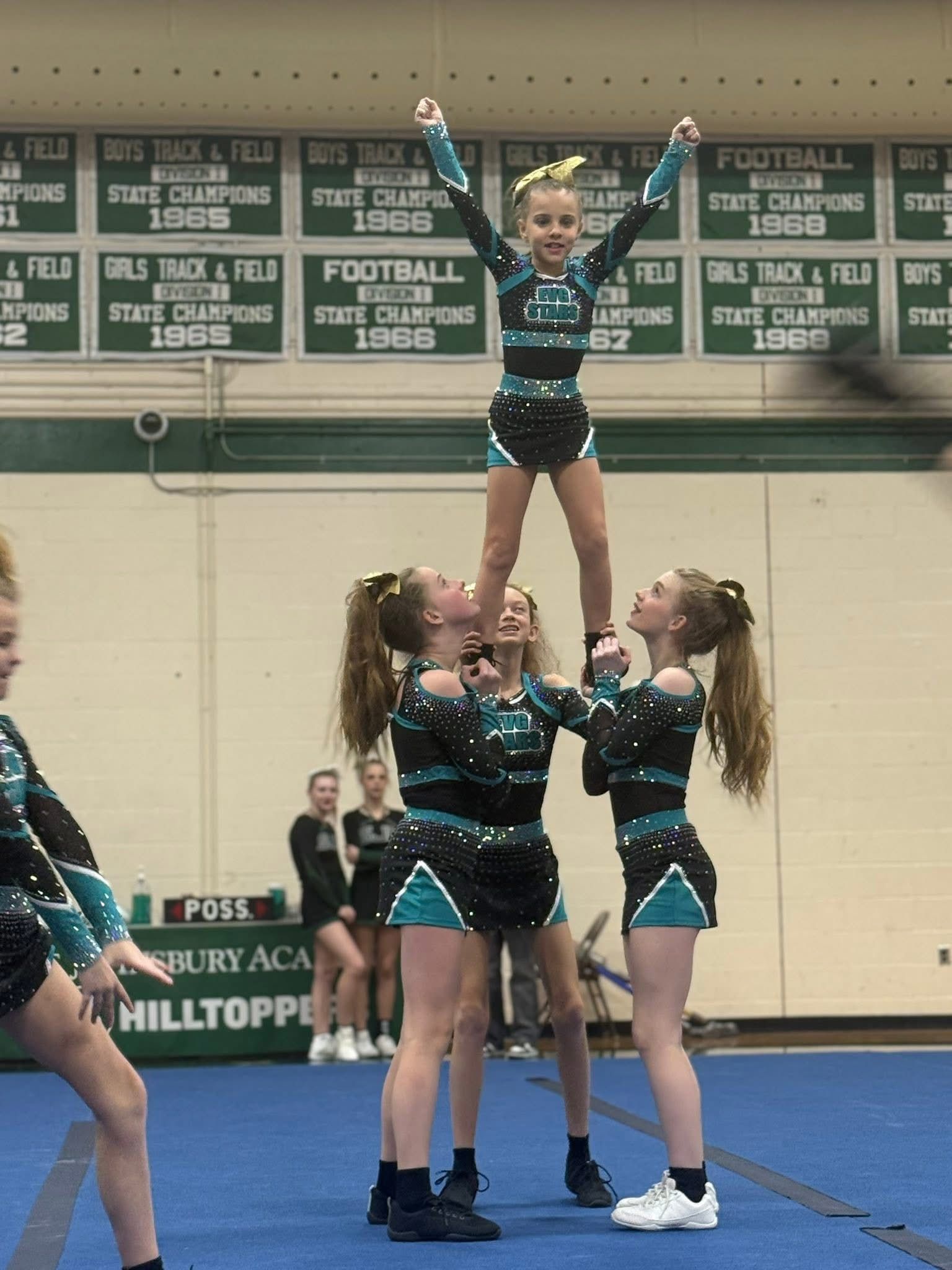 Cheerleaders in blue and black uniforms perform a stunt in a gymnasium, lifting one athlete overhead.