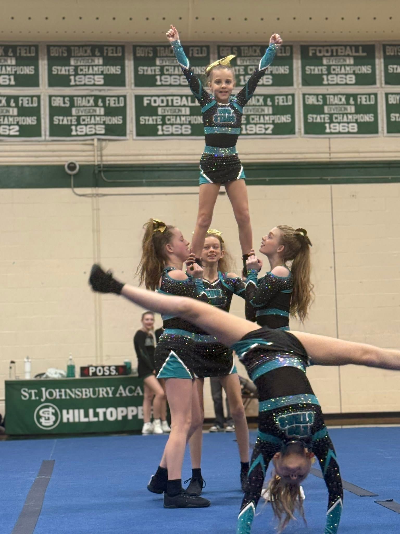 Cheerleaders perform a stunt in a gym, one flyer held overhead on a blue mat.