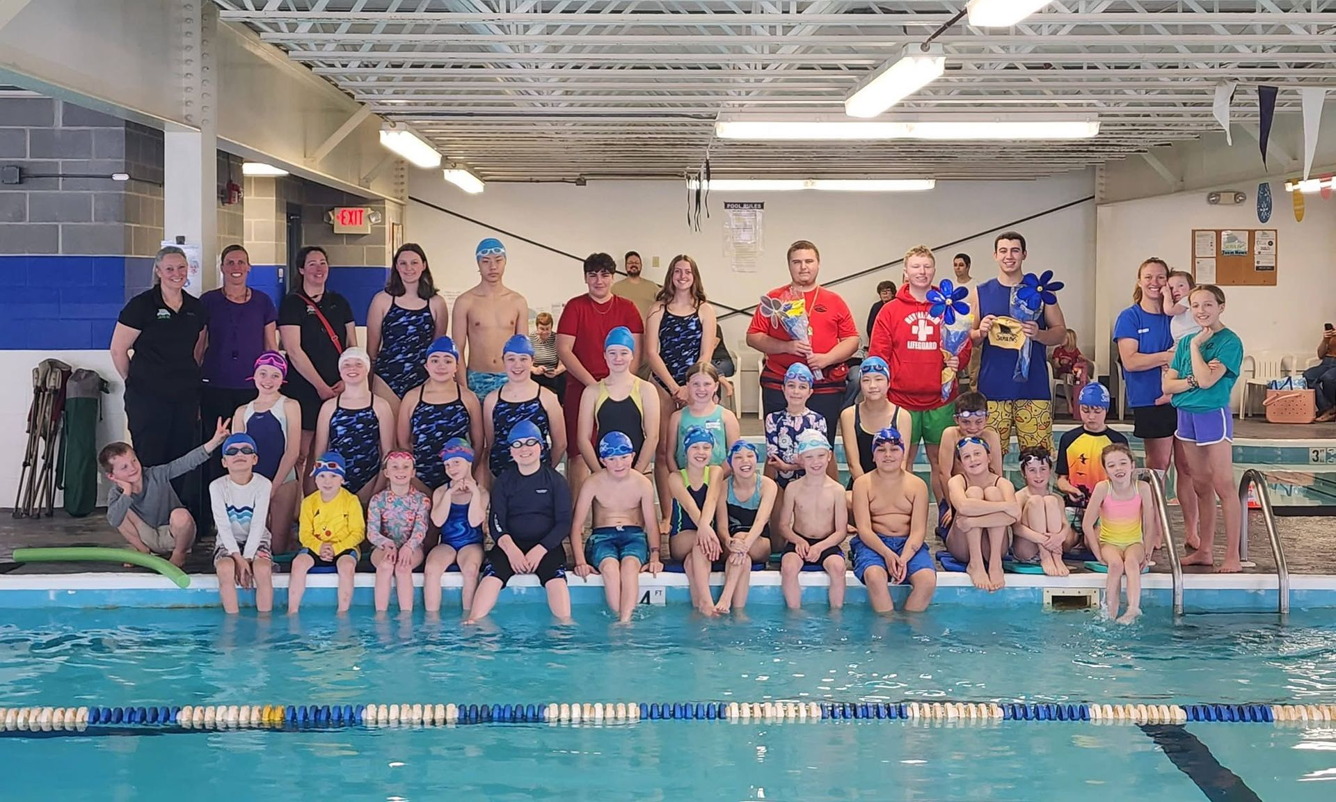 Group of swimmers posing on pool deck with medals, in front of indoor swimming pool lanes