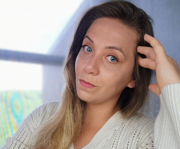 Woman with brown-and-blonde hair resting her hand on her head, wearing a white sweater indoors.
