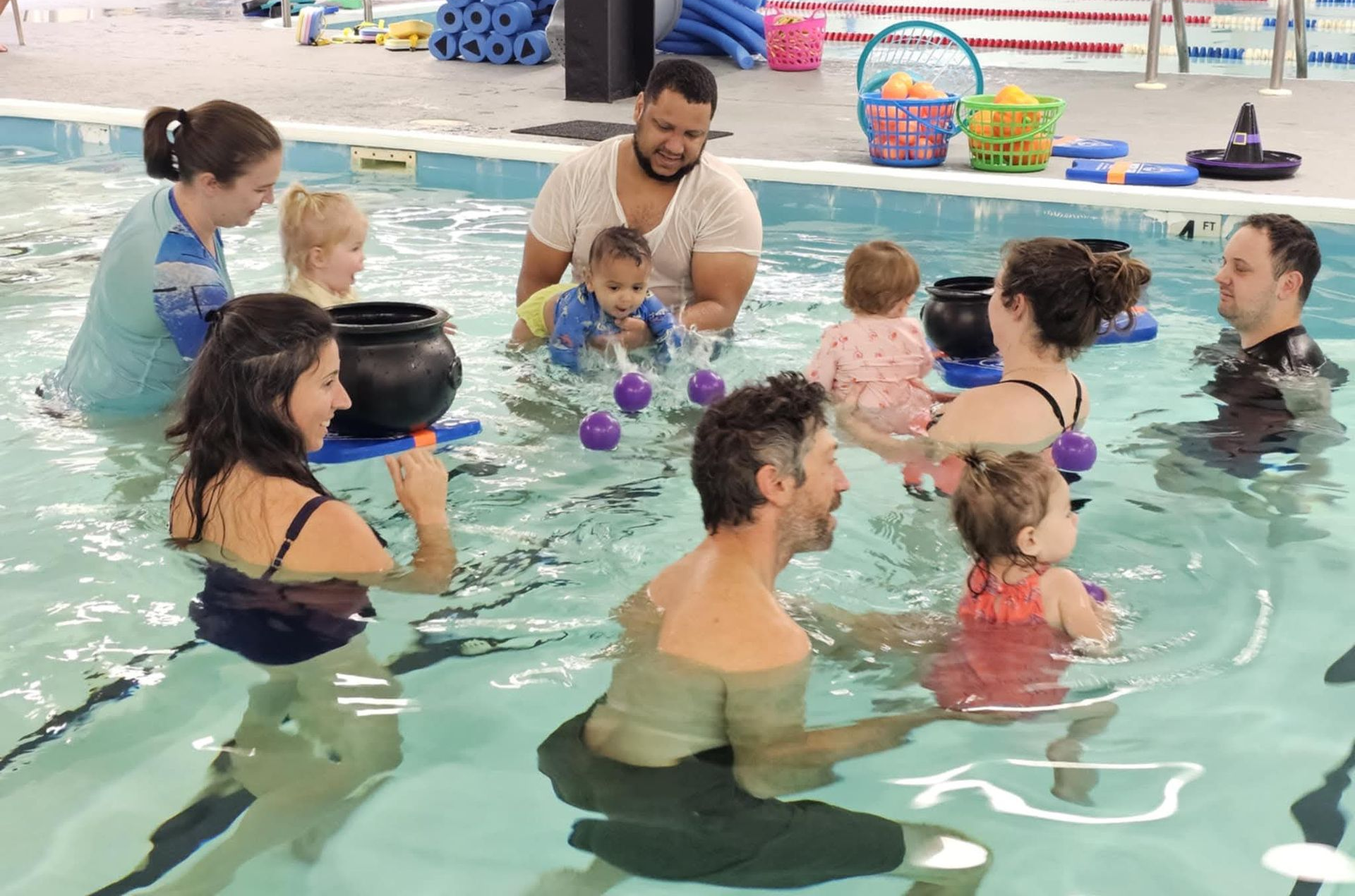 Adults and children in a swimming pool during a group water lesson, with colorful toys.