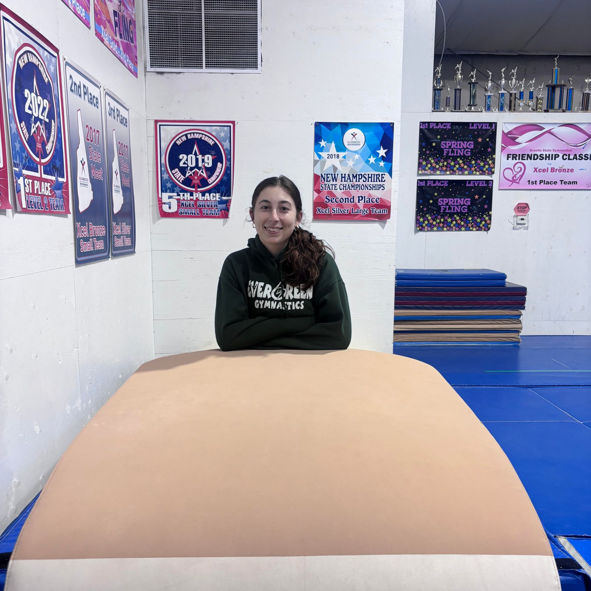 Smiling person in a gymnastics gym behind an orange mat, with posters on the wall.