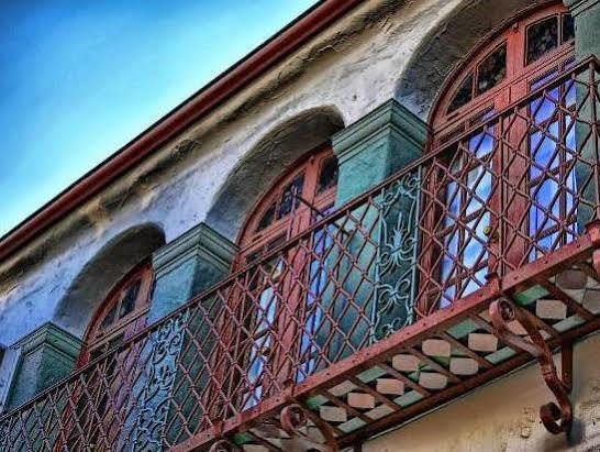 Balcony with rust-colored ironwork, arched windows, and teal-colored accents on an old building with a blue sky.