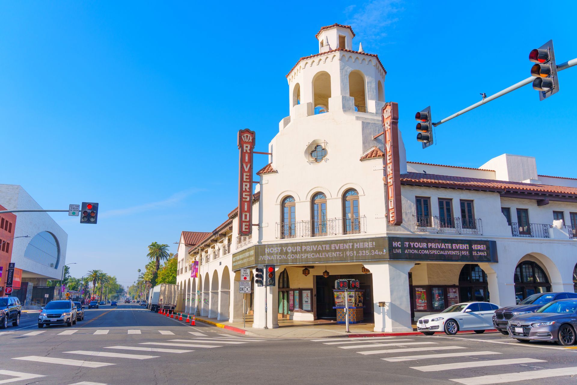 White stucco theater building with tower on a sunny street, cars, and traffic lights.
