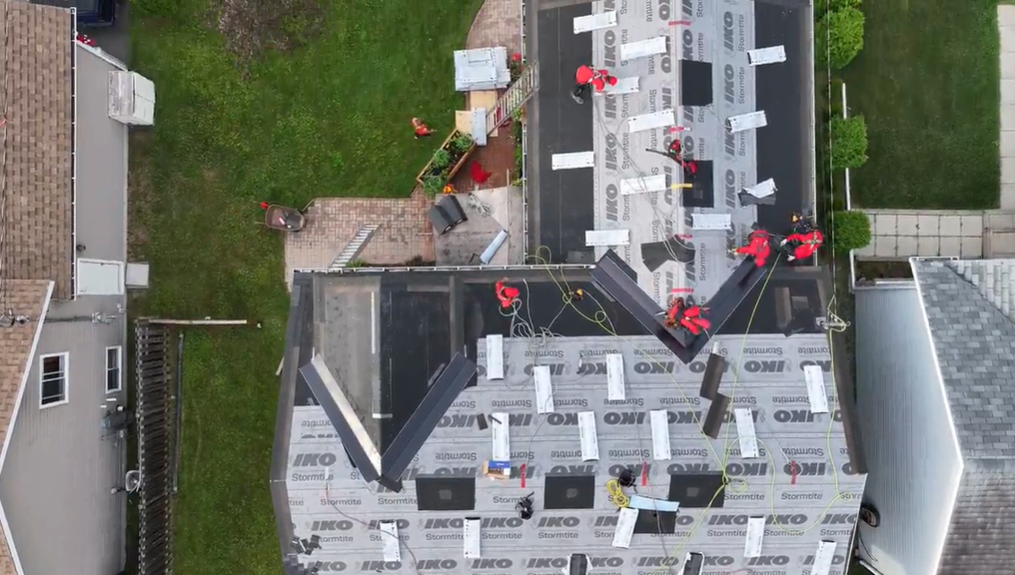Aerial view of workers installing roofing on a house next to a yard and driveway.