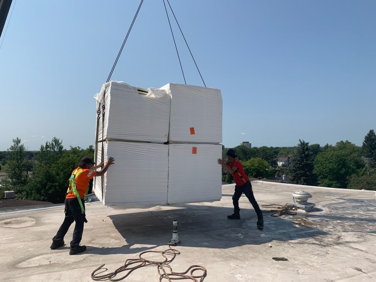 Two workers guide a suspended stack of white insulation blocks on a rooftop under a blue sky.